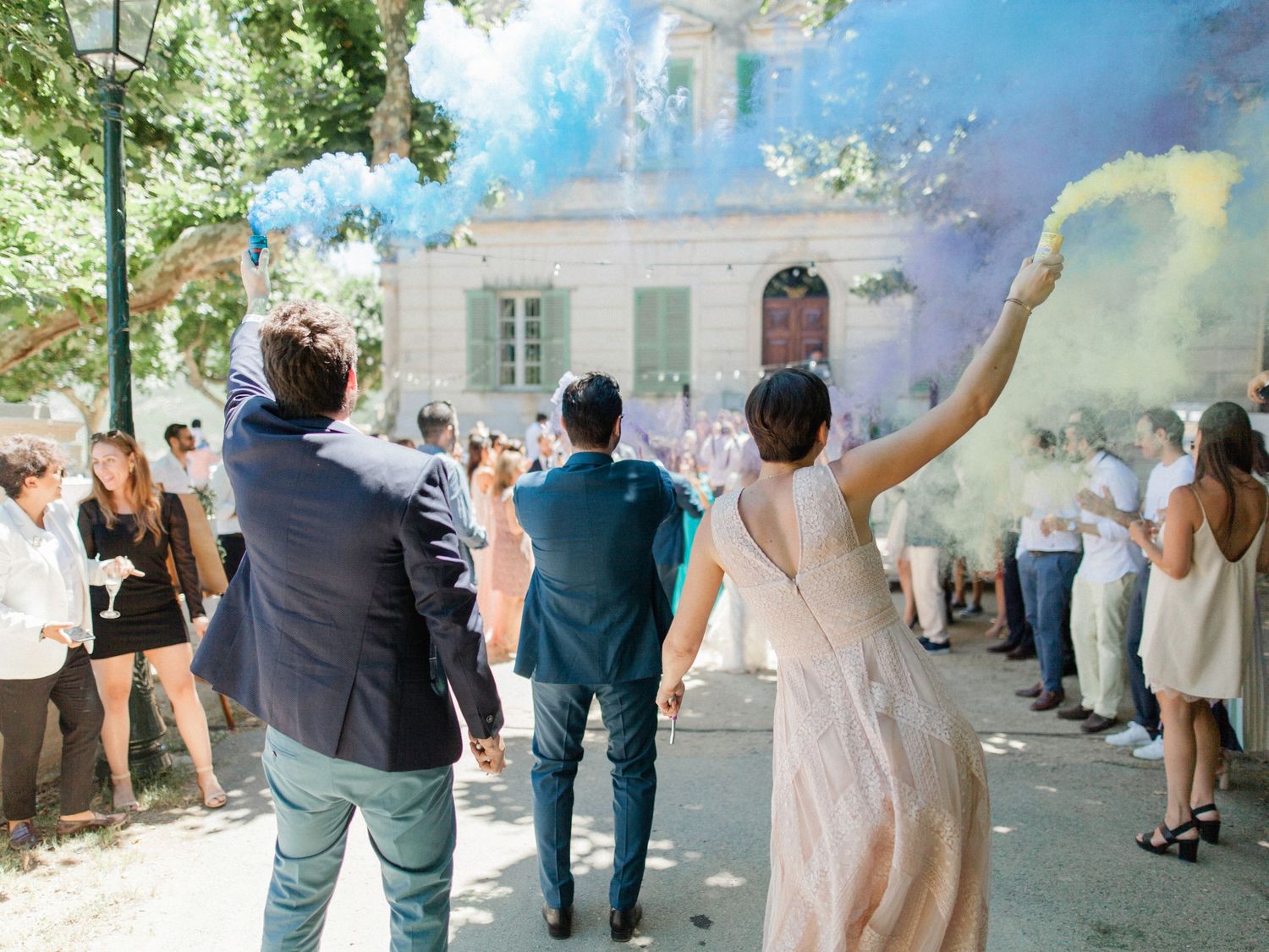 Wedding guests celebrating outdoors with colorful blue smoke bombs, walking toward a charming white venue surrounded by trees.