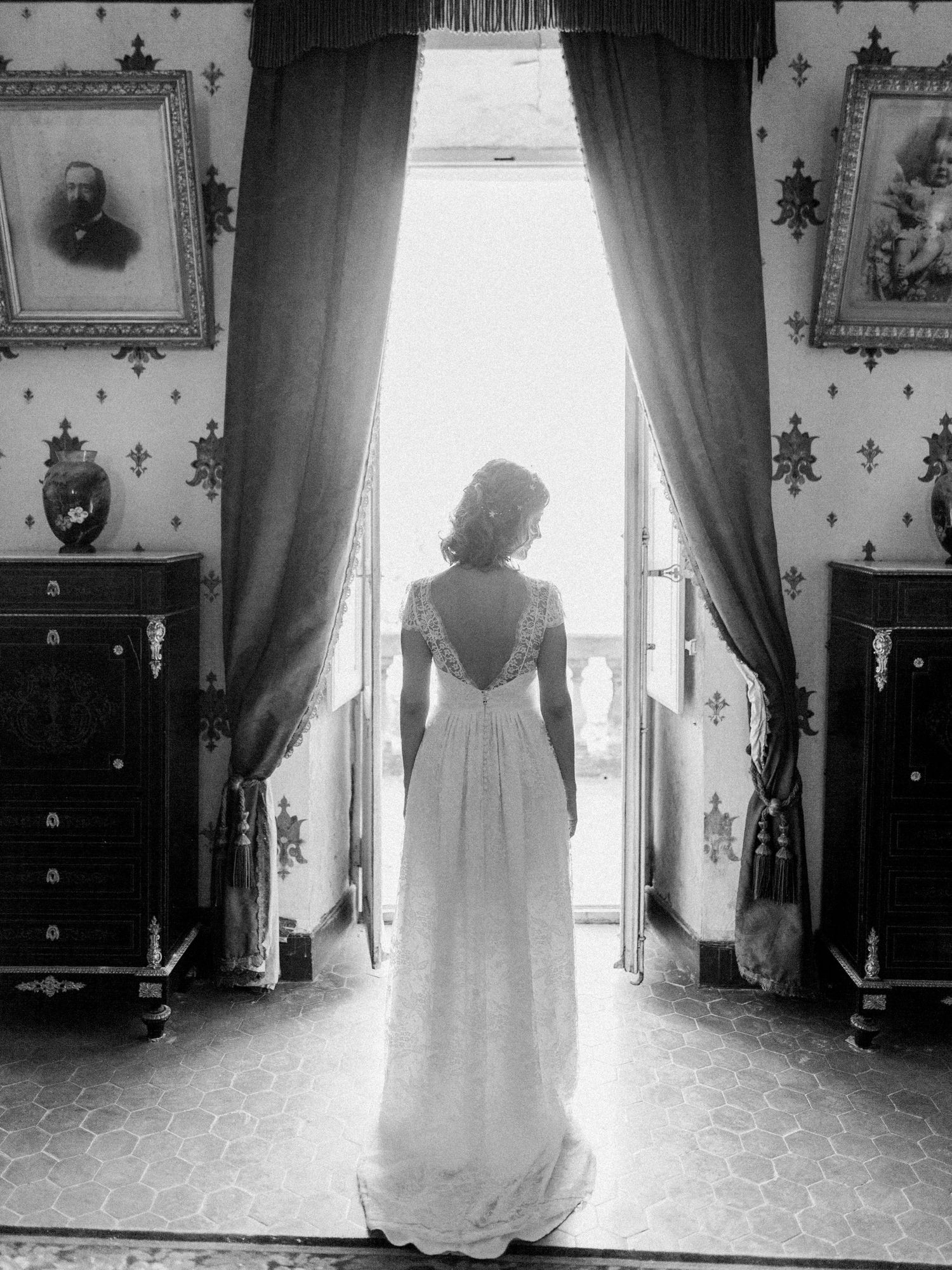 Black and white photo of a bride in a lace gown standing at an open window in an elegant vintage room.
