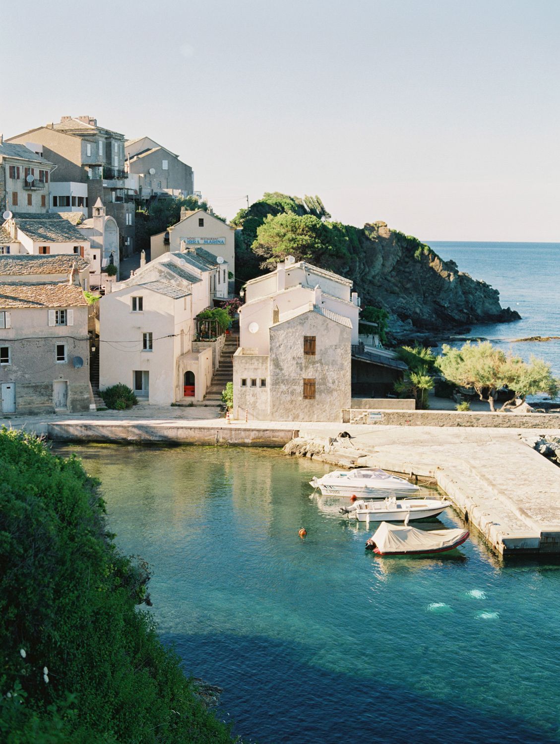 Charming coastal village with stone buildings, crystal-clear turquoise water, and small boats docked at a quiet Mediterranean harbor.