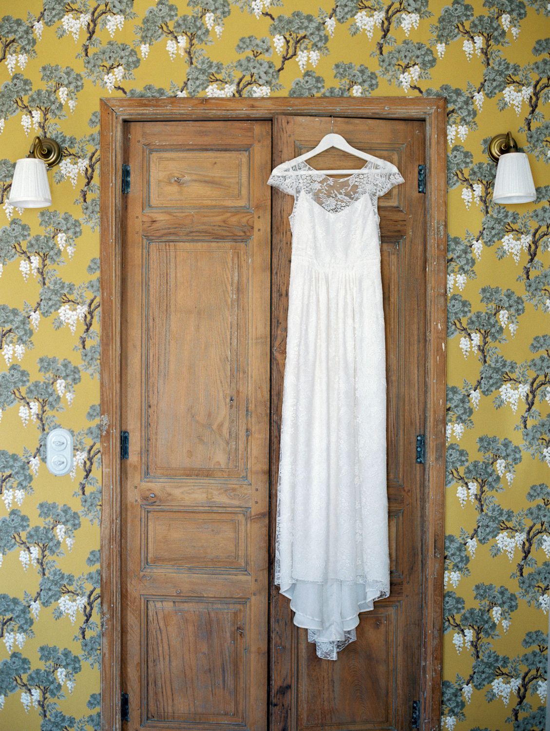 White lace wedding dress hanging on wooden double doors against yellow floral wallpaper with wall sconces.