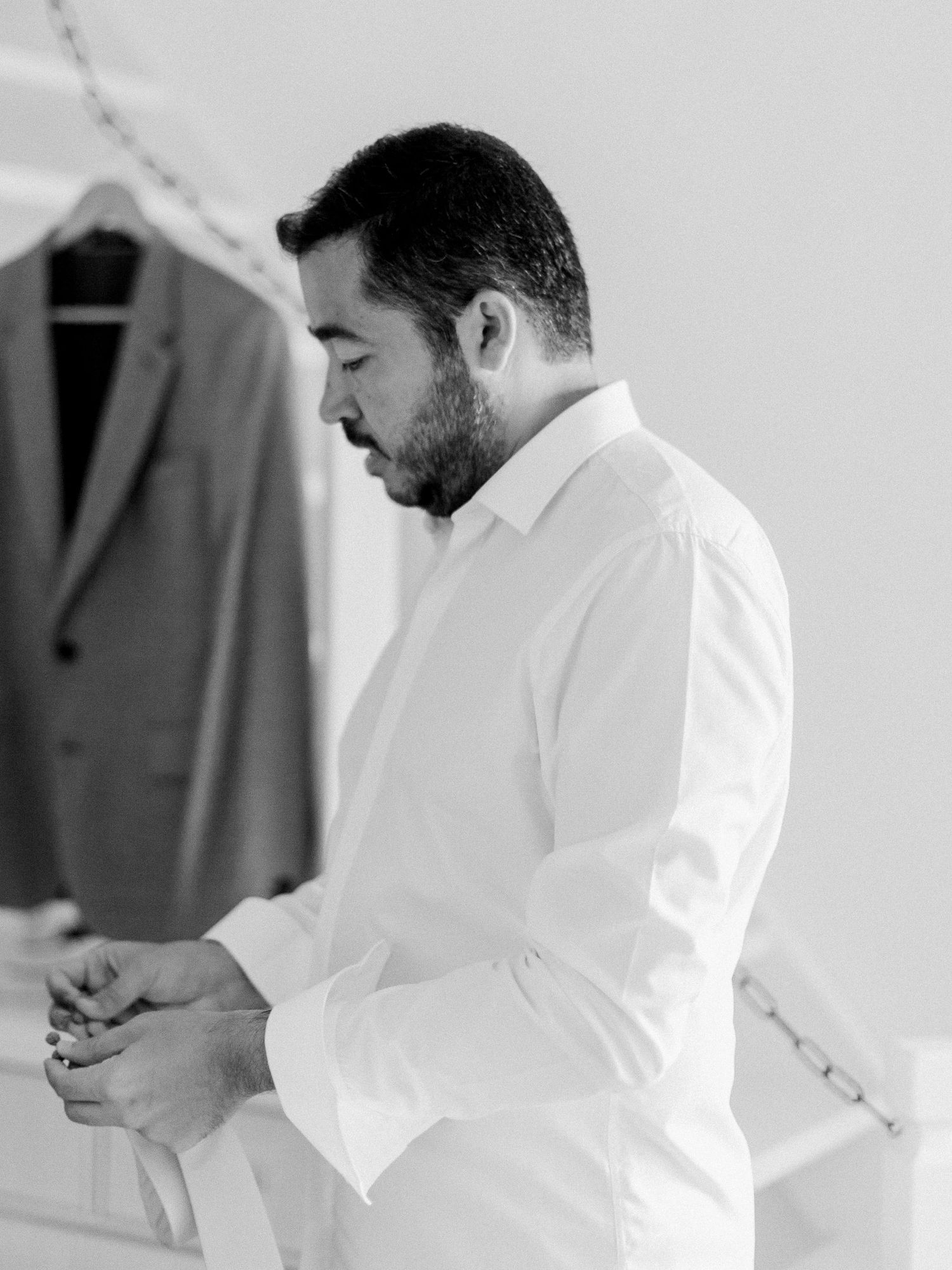 Photo en noir et blanc d'un marié barbu, vêtu d'une chemise blanche, boutonnant ses poignets avant la cérémonie de mariage.