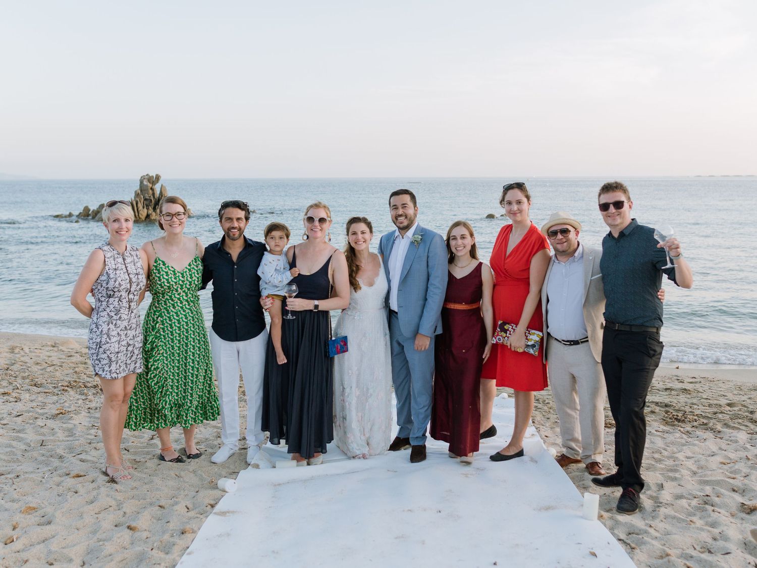 Groupe d'invités de mariage posant ensemble sur une plage de sable au coucher du soleil, avec l'océan en arrière-plan.