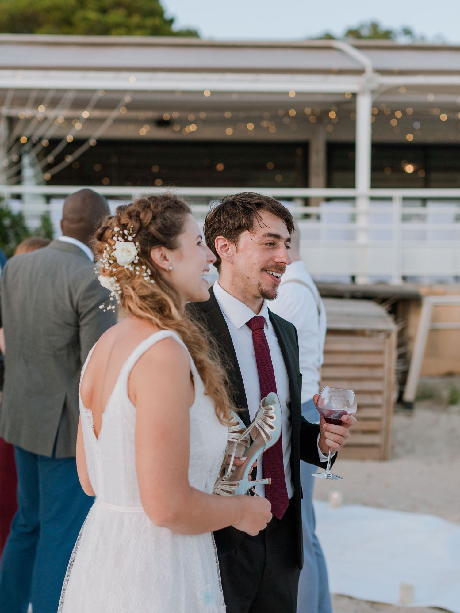 Les mariés sourient en tenant des verres de vin lors d'une réception de mariage en plein air sur la plage, décorée de guirlandes lumineuses.
