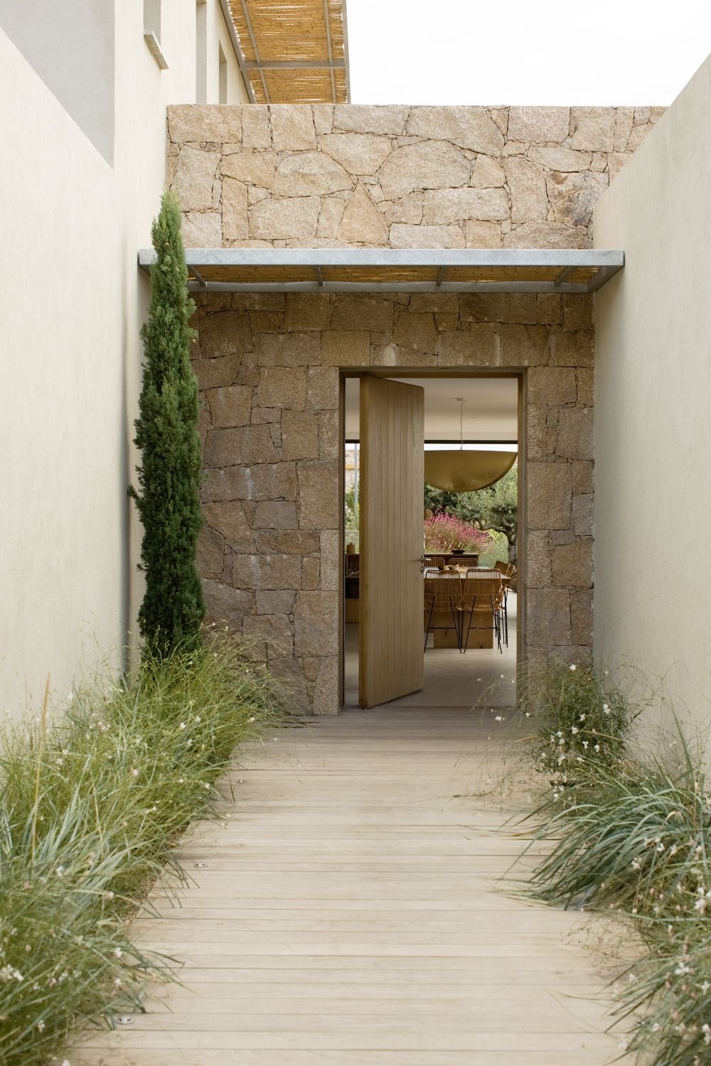 Entrée de la villa aux murs de pierre, avec allée en bois, cyprès et vue sur la salle à manger à travers la porte ouverte à porto-vecchio