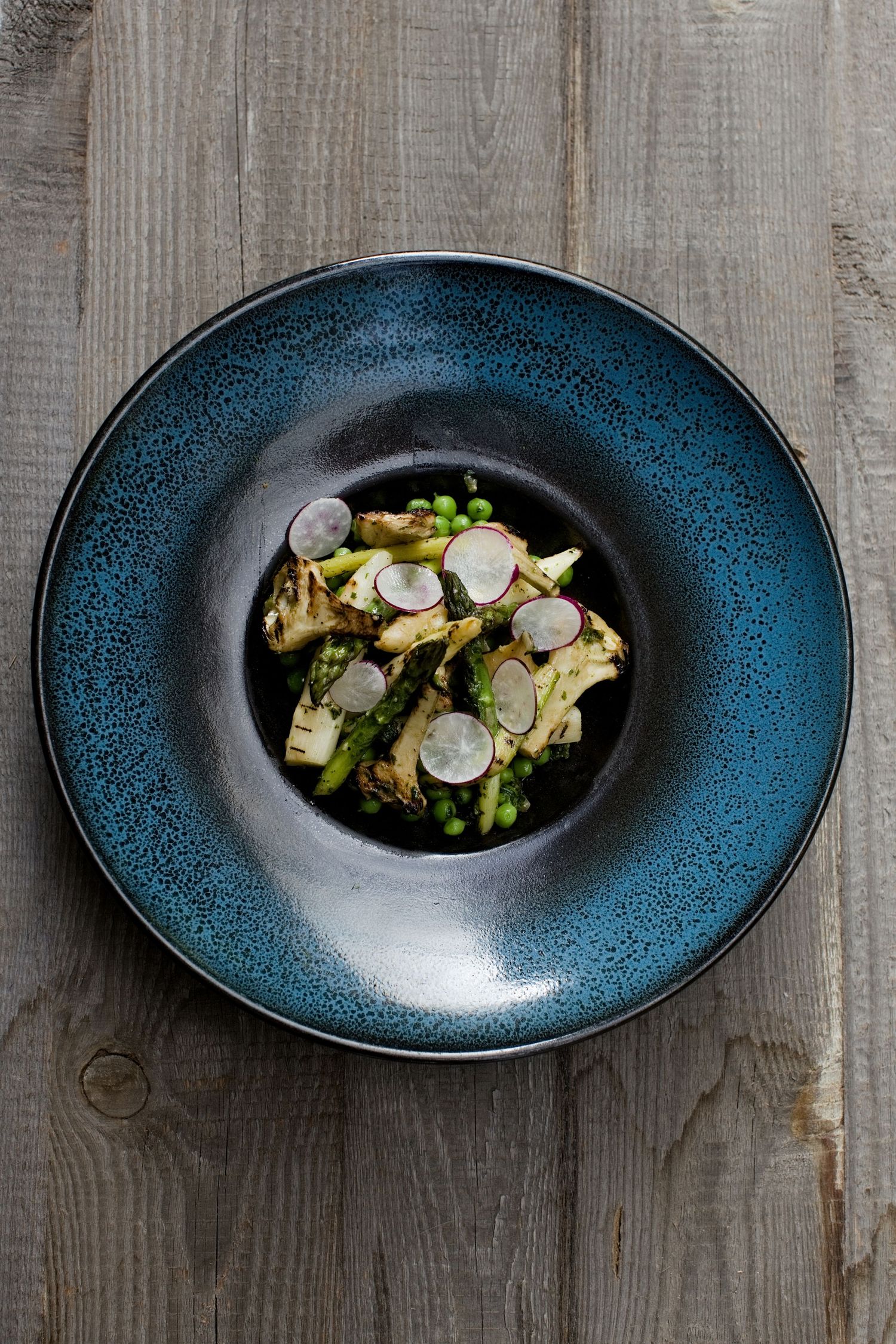Plat artistique d'artichauts grillés, de petits pois et de radis tranchés, servi dans un bol en céramique bleue.