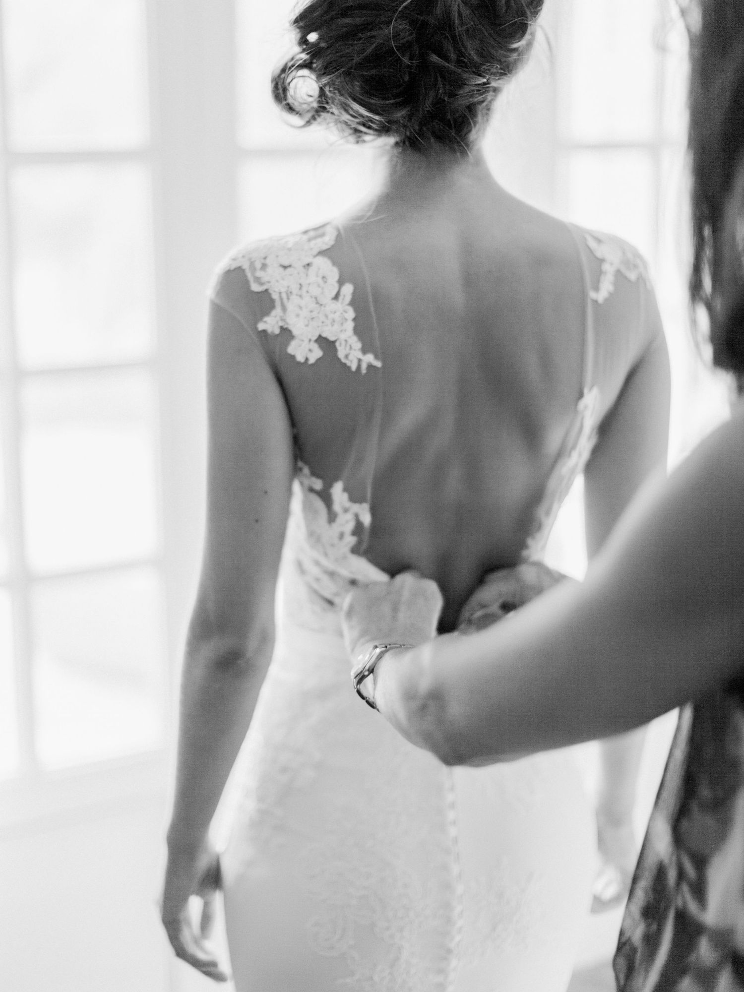 Photo en noir et blanc d'une mariée en robe de mariée dos nu en dentelle, boutonnée par une demoiselle d'honneur.