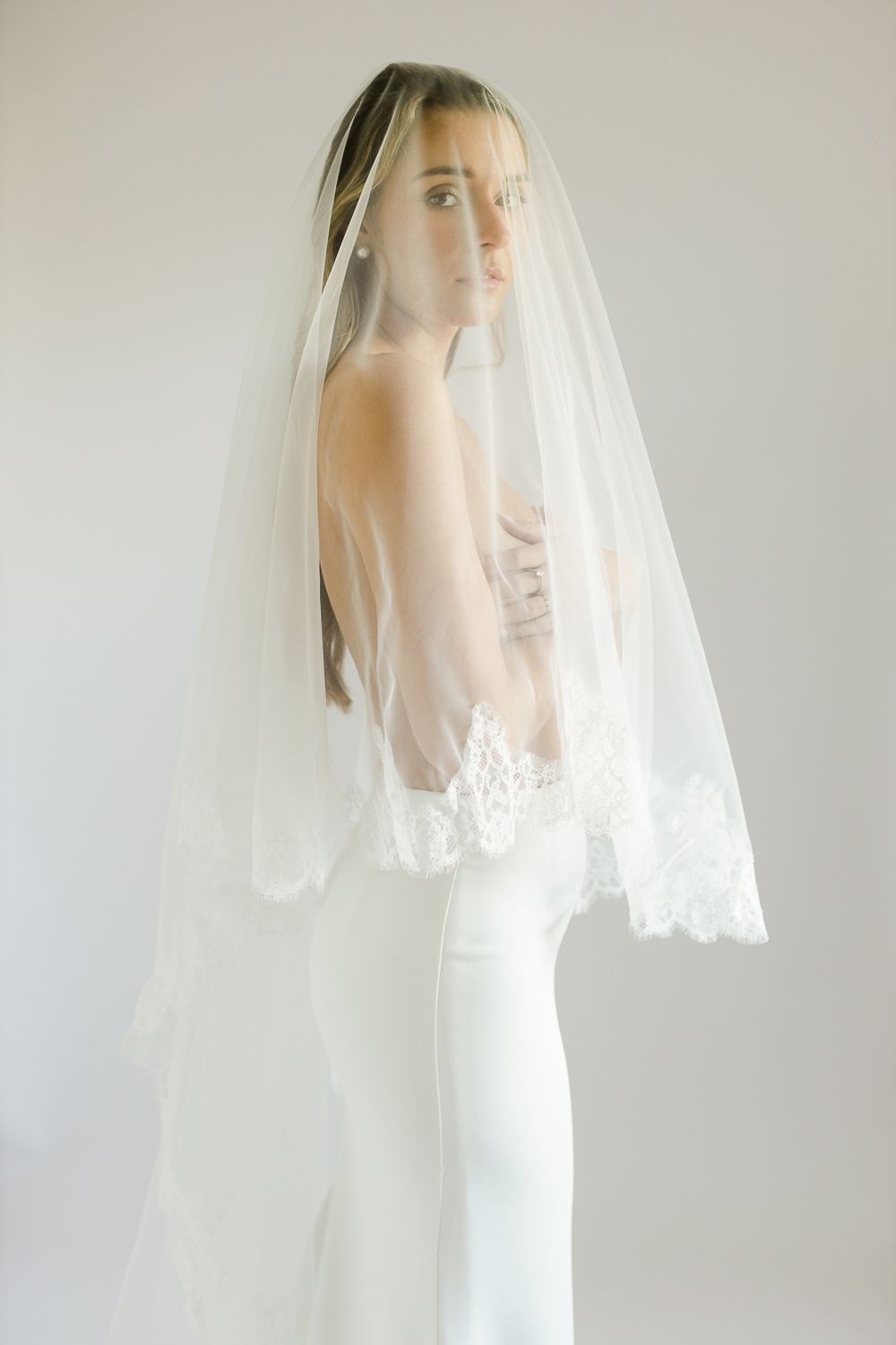 Bride wearing a white lace-trimmed wedding veil over a strapless gown, posing elegantly against a light background.