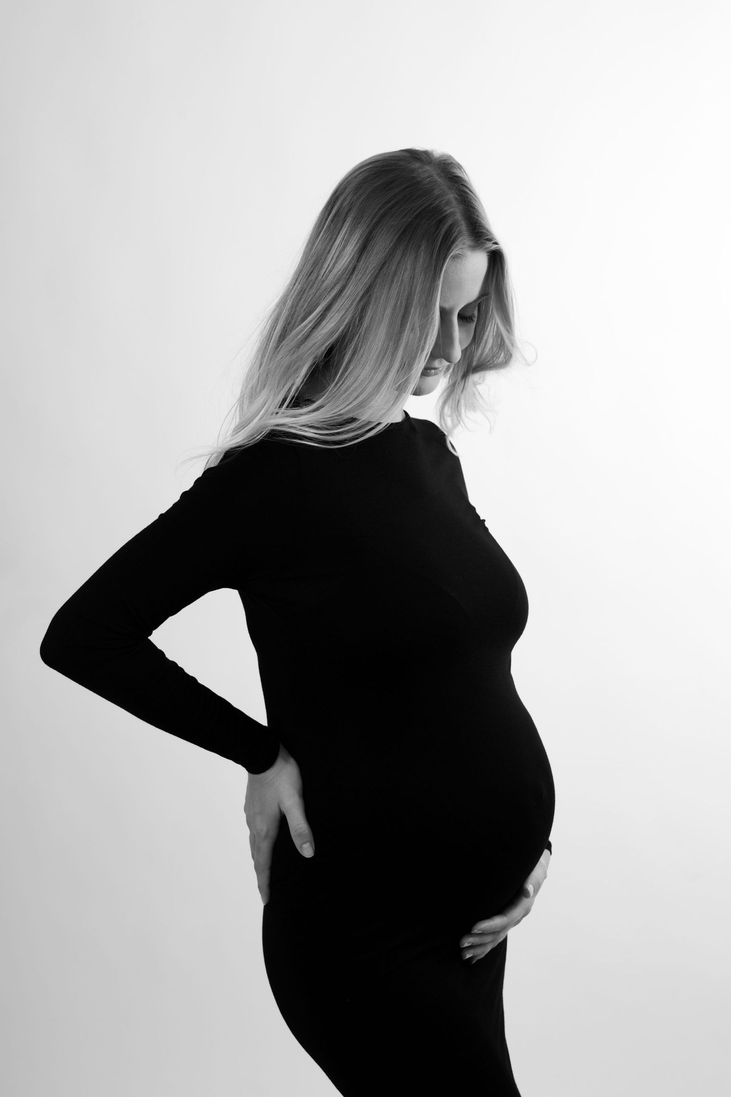 Profil latéral en noir et blanc d'une femme enceinte en robe noire, tenant délicatement son ventre arrondi.