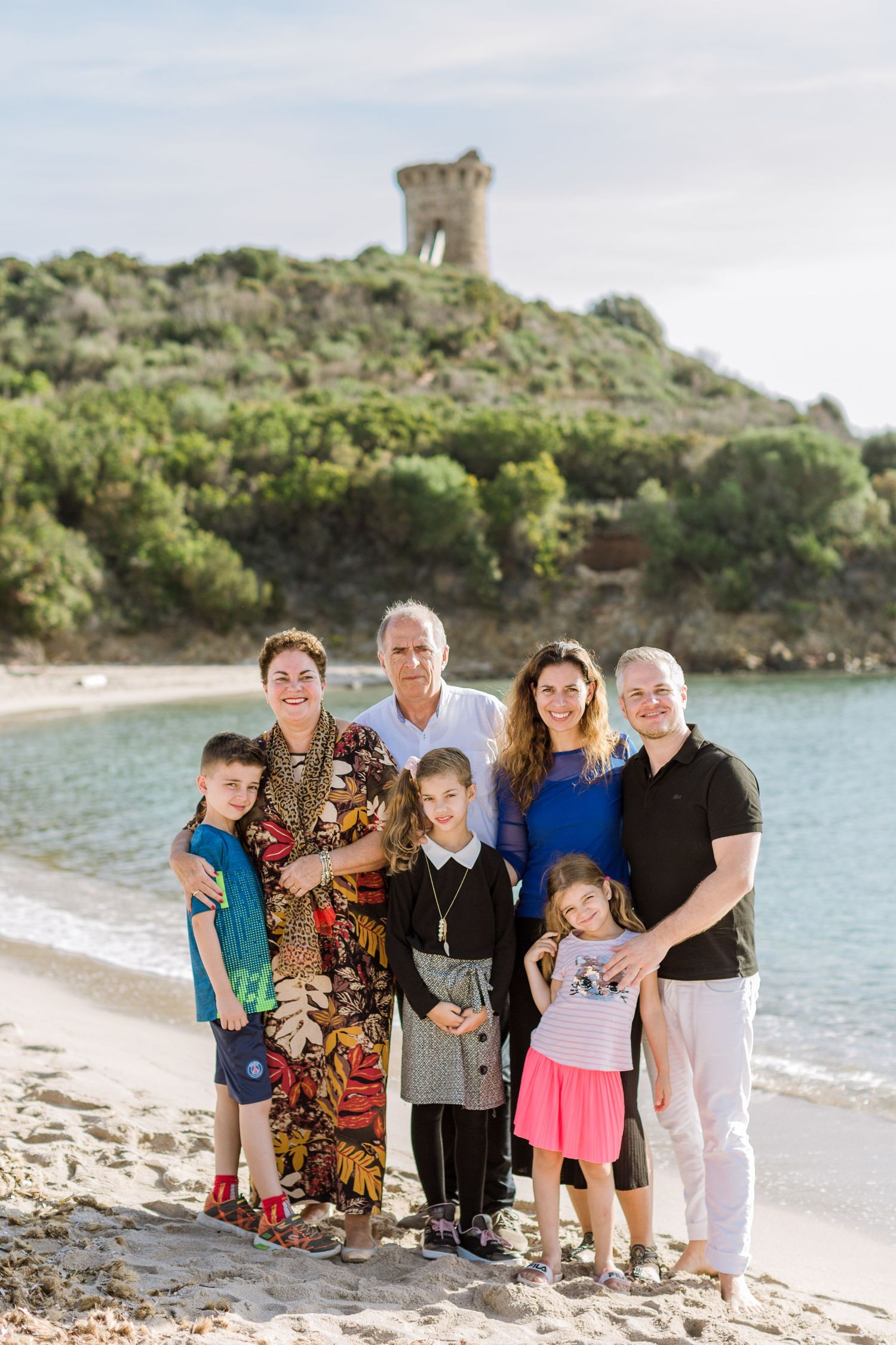 Une famille multigénérationnelle pose ensemble sur la plage de Fautea, avec une ancienne tour en pierre sur une colline en arrière-plan.