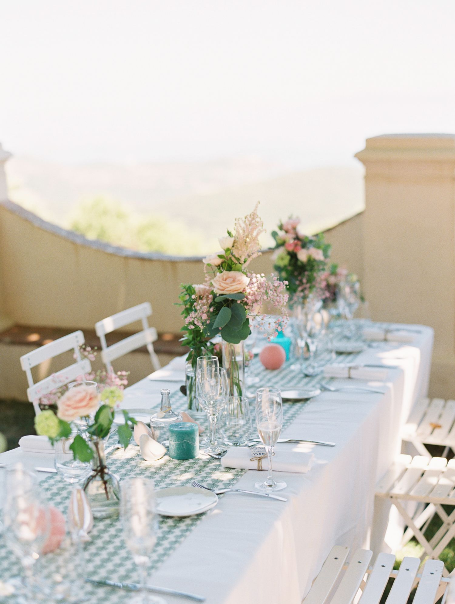 Elegant outdoor wedding reception table with floral centerpieces, glassware, and soft blue linens against a scenic backdrop.