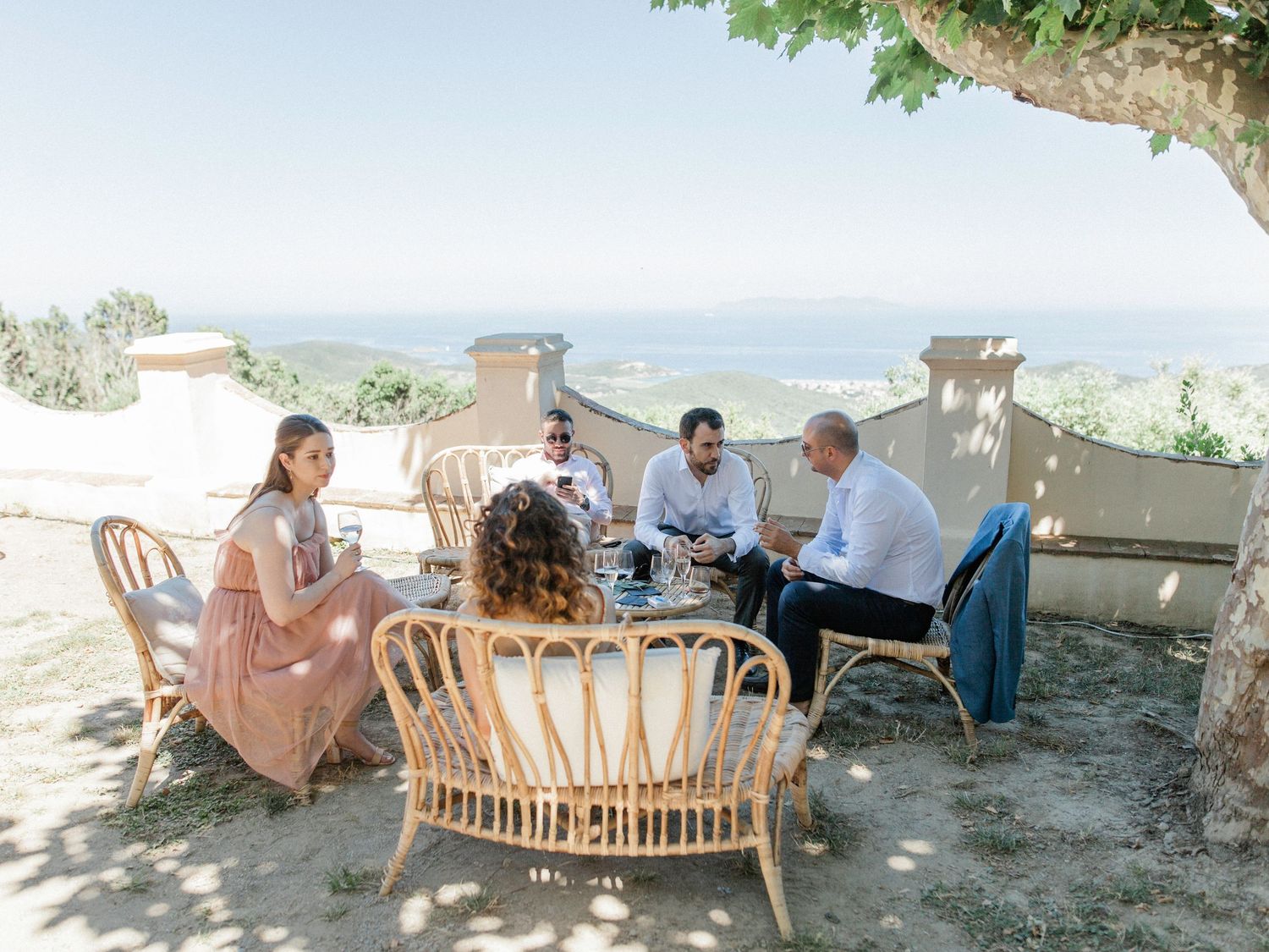 Four people dining al fresco at a rustic wicker table with scenic Mediterranean hillside views on a sunny day.