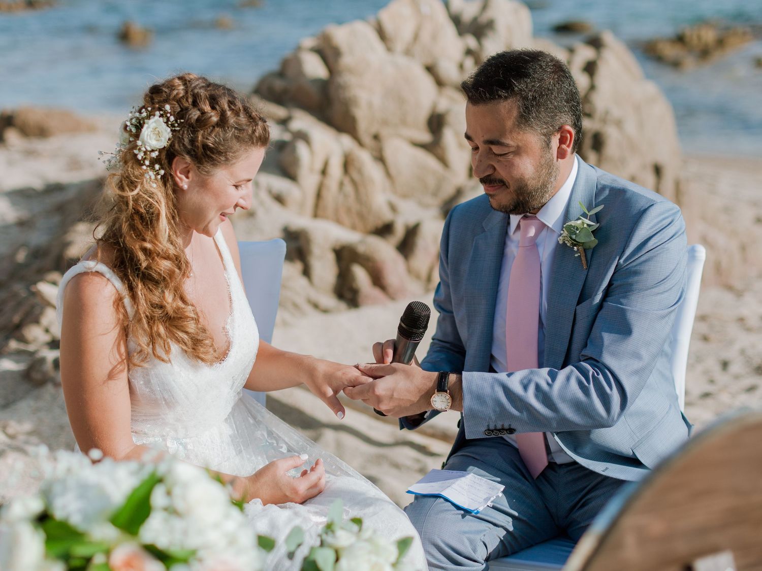 Le marié passe l'alliance au doigt de la mariée lors d'une cérémonie romantique sur la plage, avec pour toile de fond une côte rocheuse.