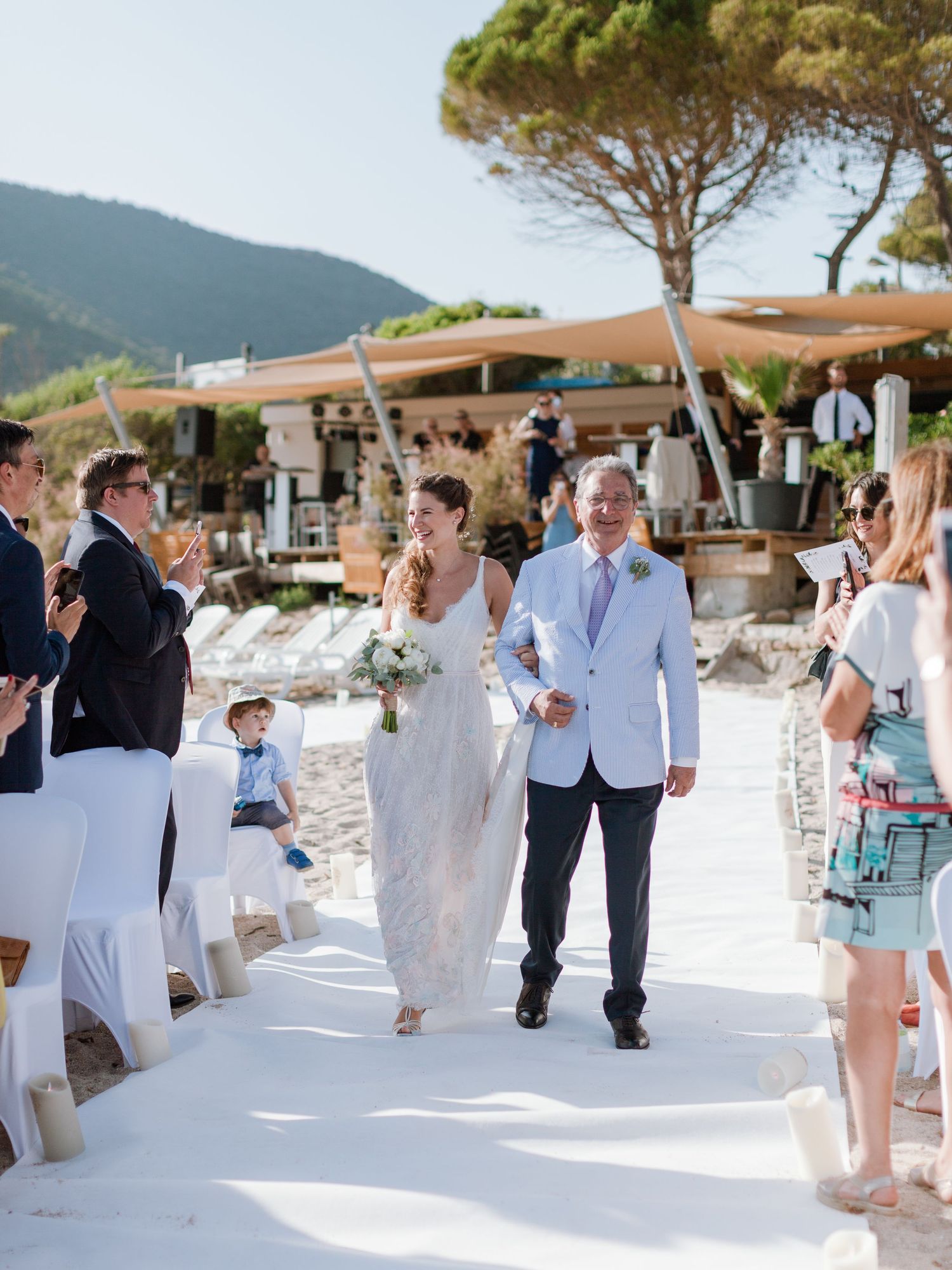 Une mariée s'avance dans l'allée blanche, accompagnée, lors d'une cérémonie de mariage en plein air sur la plage ; les invités applaudissent