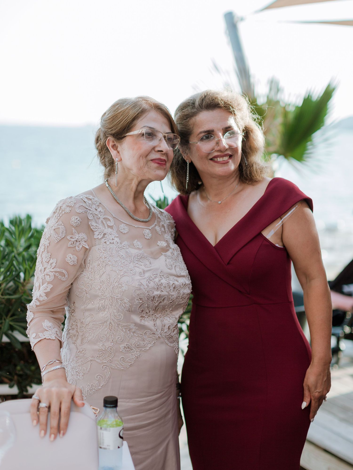 Deux femmes élégamment vêtues posant ensemble lors d'un événement en plein air près de la mer, l'une en dentelle beige, l'autre en bordeaux.