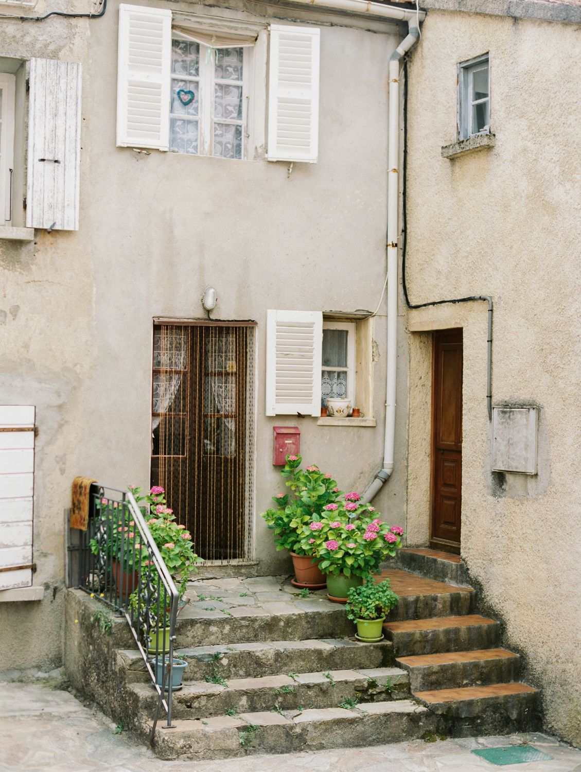 Charming French village stone building with white shutters, potted pink flowers, and stone steps leading to rustic doors.