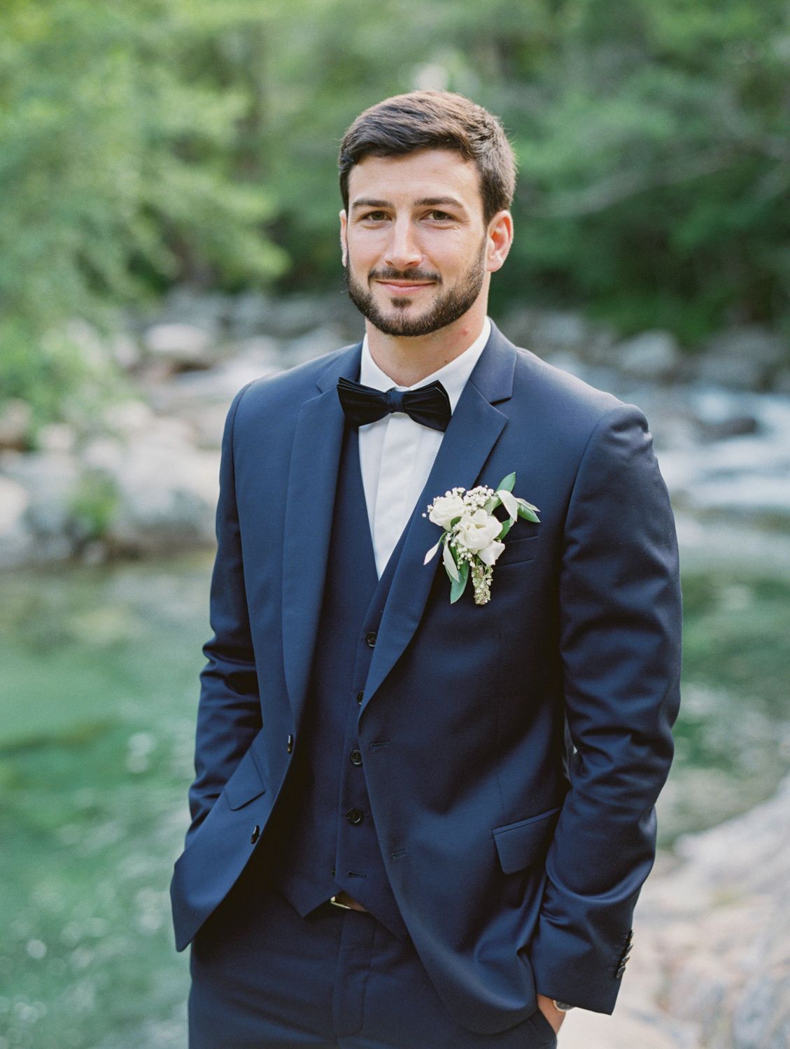A groom in a navy blue three-piece suit with bow tie and white boutonniere poses by a river.