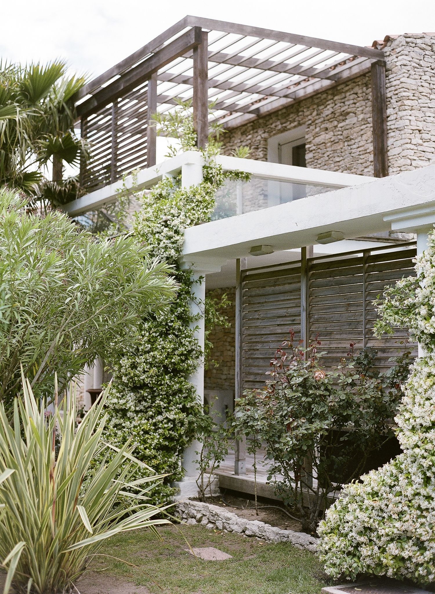 Un jardin luxuriant entoure une villa moderne en pierre avec une pergola en bois, des volets blancs et des plantes grimpantes