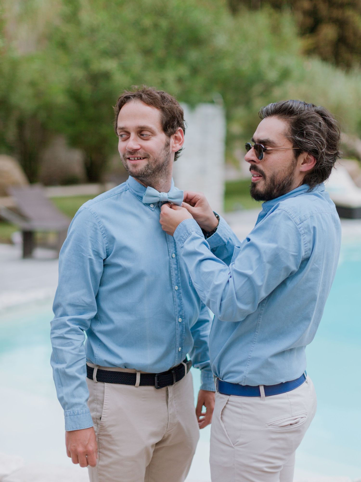 Deux hommes en chemises bleues assorties, l'un ajustant le nœud papillon de l'autre près d'une piscine lors d'un événement en plein air.