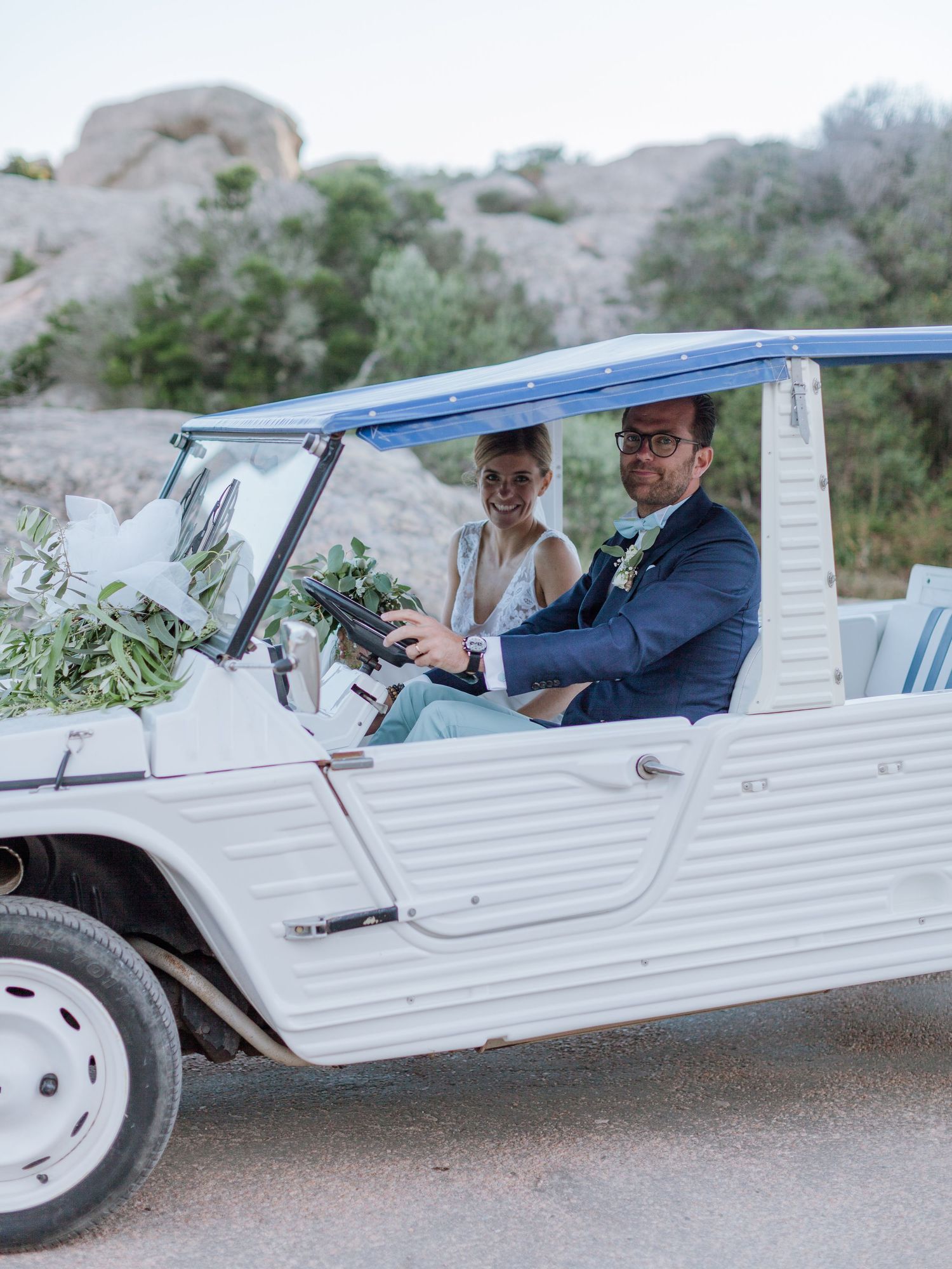 Les mariés se promènent dans une voiture vintage blanche décapotable décorée de verdure le jour de leur mariage.