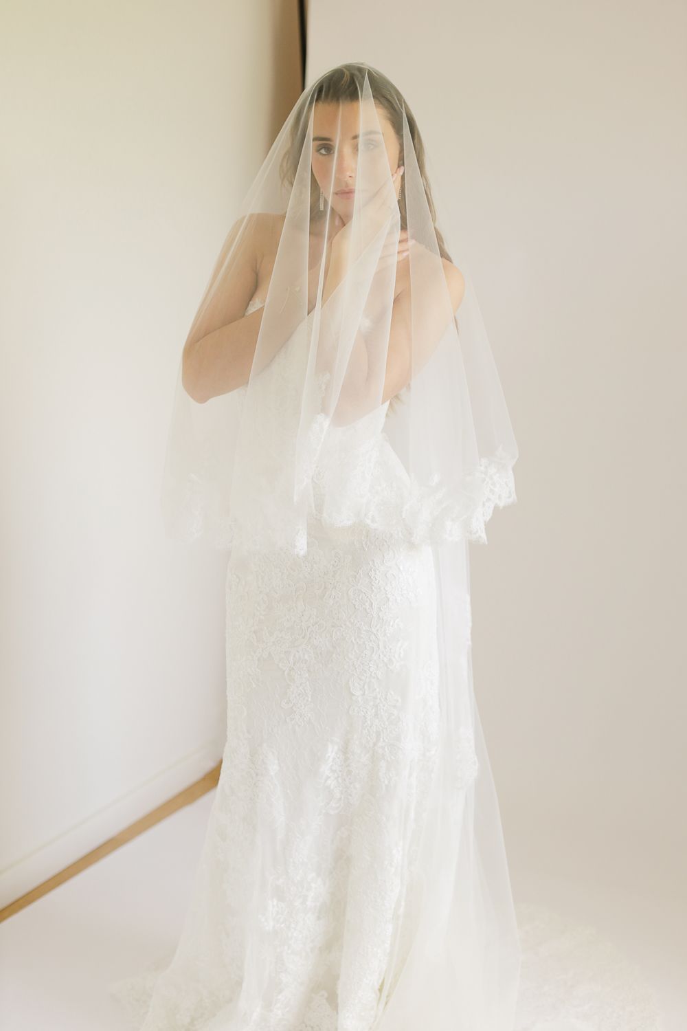 Bride in lace wedding gown with sheer veil draped over face, posing elegantly in a bright white room.