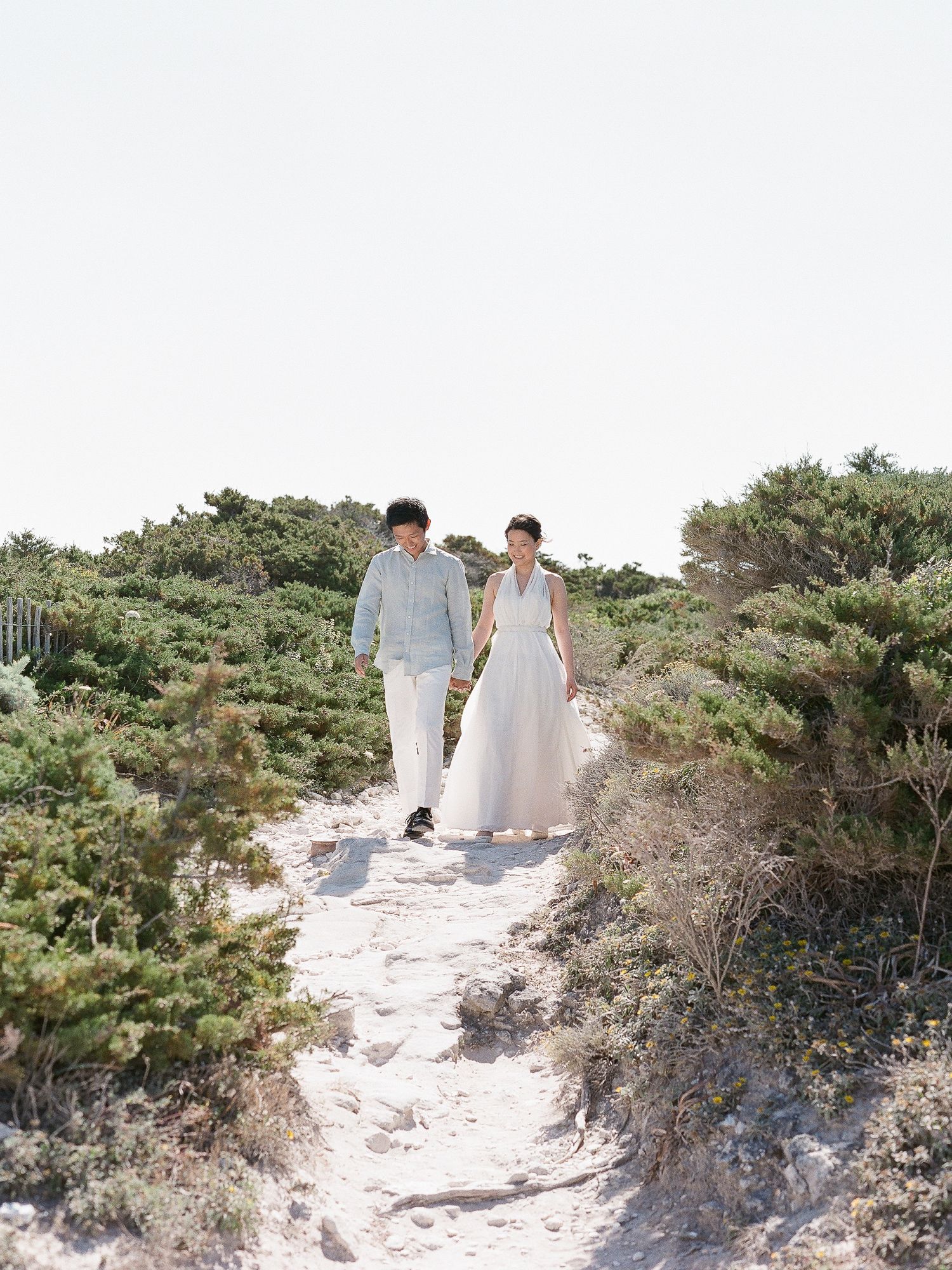 Un couple en tenue de mariage se tient la main en marchant le long d'un sentier côtier sablonneux entouré de buissons verdoyants.