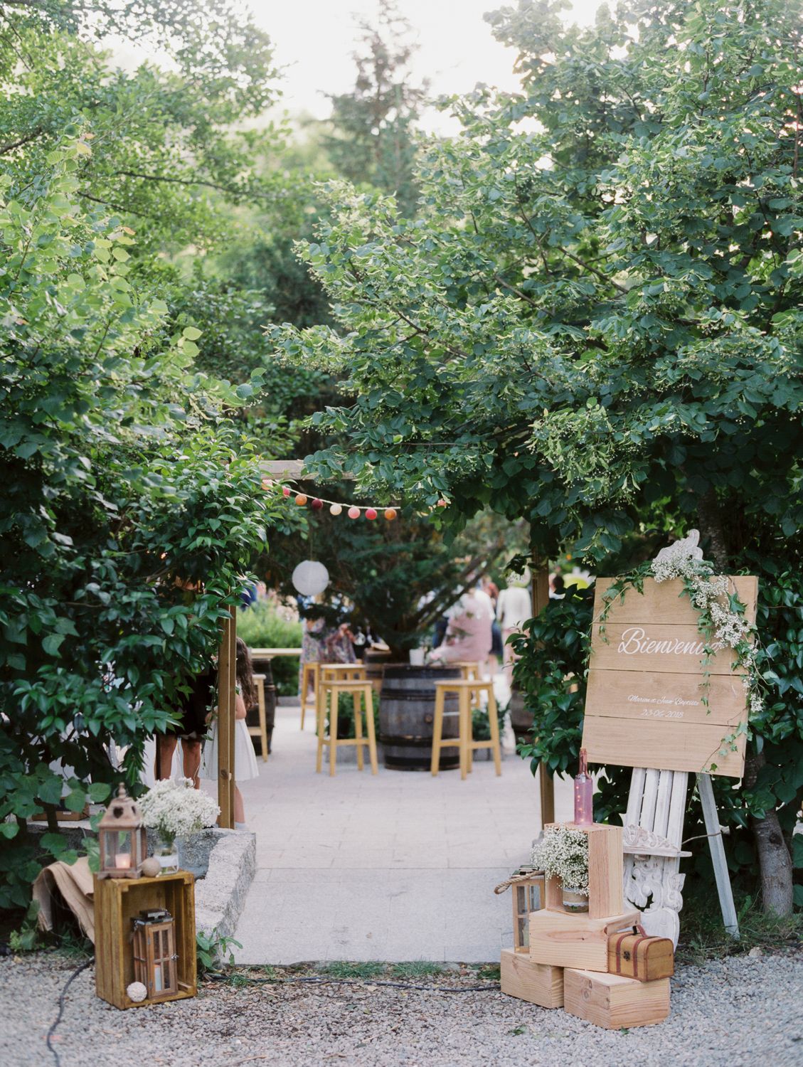 Rustic outdoor wedding venue entrance with wooden crates, welcome sign, string lights, and lush green trees lining a garden path.