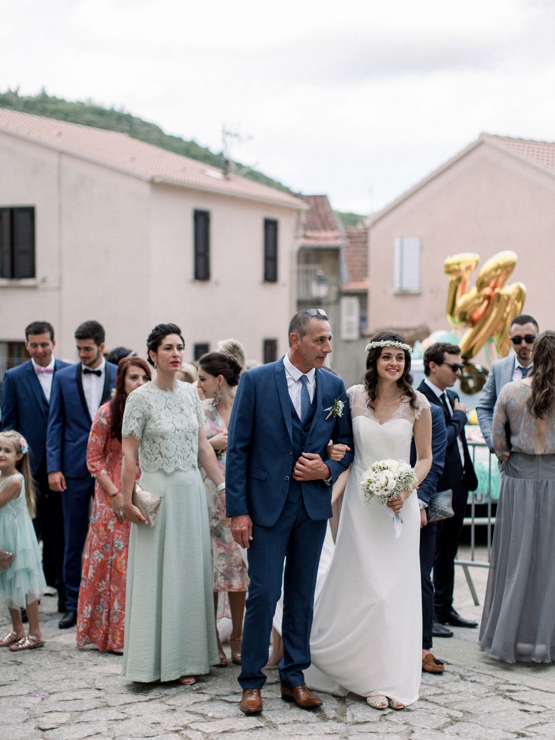 Bride and groom walk with wedding guests in a European village square, bride holding white bouquet in elegant gown.