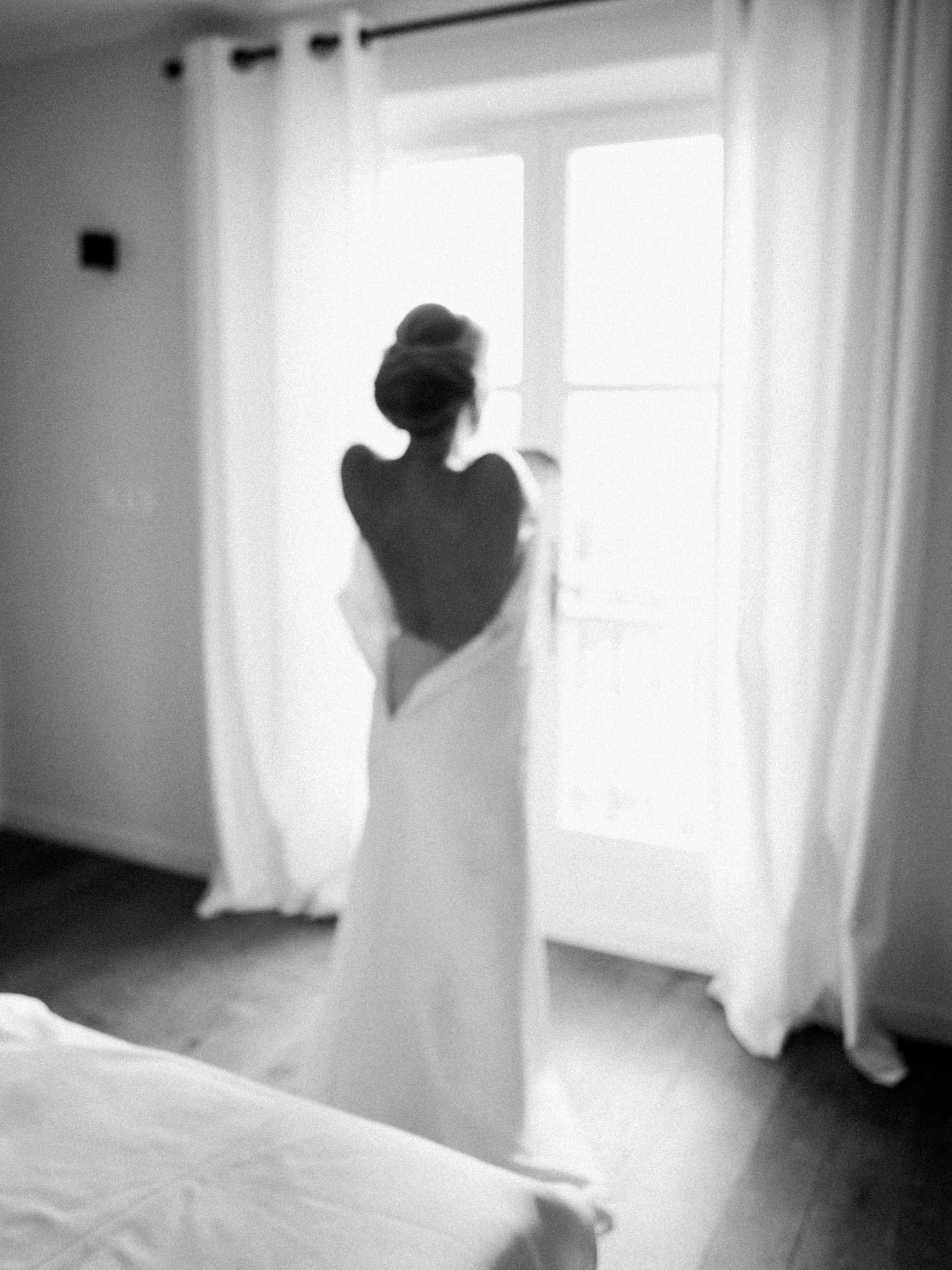 Photo en noir et blanc d'une mariée en robe de mariée dos nu, debout devant une fenêtre lumineuse aux rideaux blancs à saint Tropez.