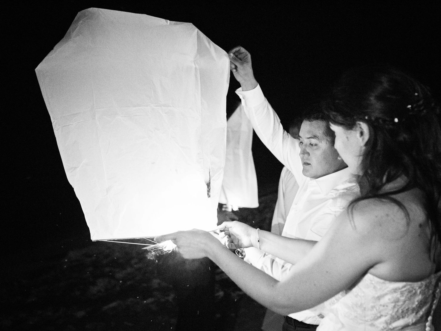 Couple en tenue de mariage lâchant une lanterne céleste ensemble la nuit, photo en noir et blanc.