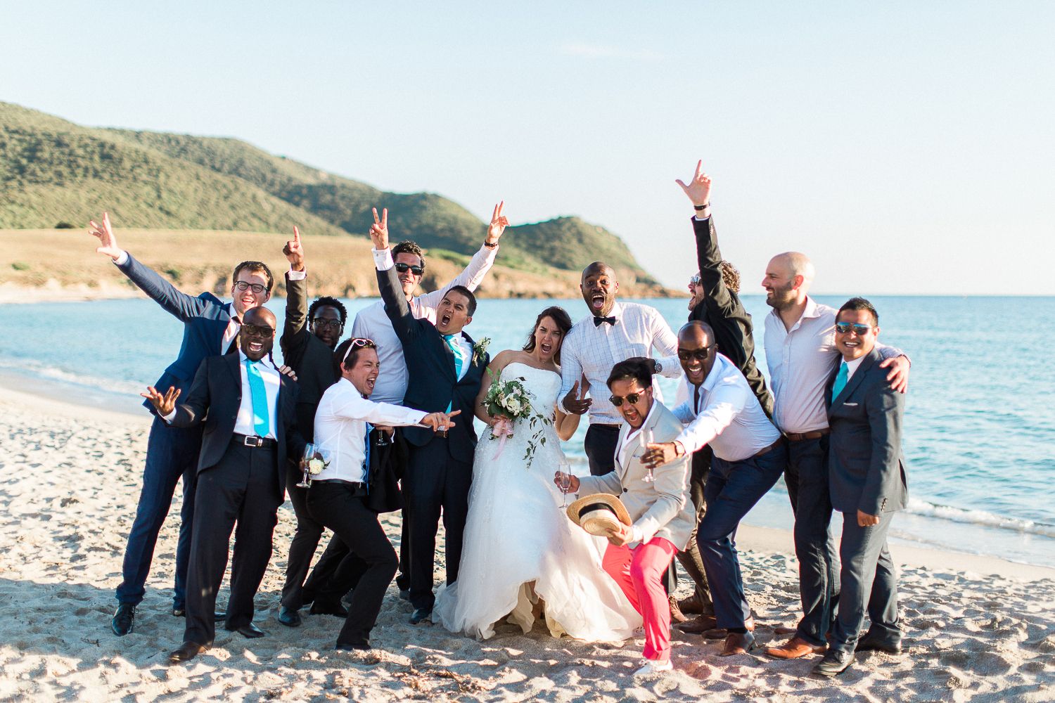 Fête de mariage sur une plage ensoleillée, les mariés entourés d'invités joyeux en tenue de soirée, avec les montagnes en arrière-plan