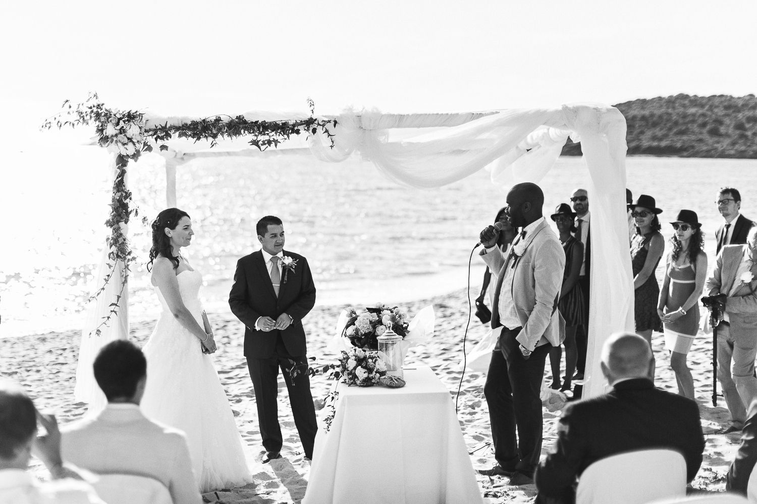 Cérémonie de mariage en noir et blanc sur la plage, avec les mariés sous une arche fleurie, les invités assis sur le rivage sablonneux.