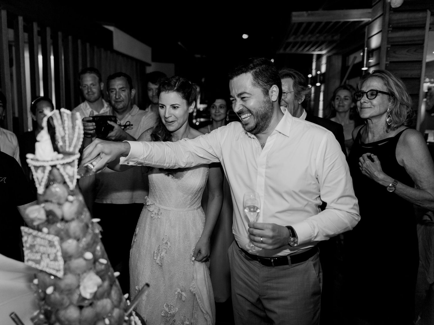 Photo en noir et blanc d'un couple coupant sa pièce montée, entouré d'invités lors d'une élégante réception en soirée.
