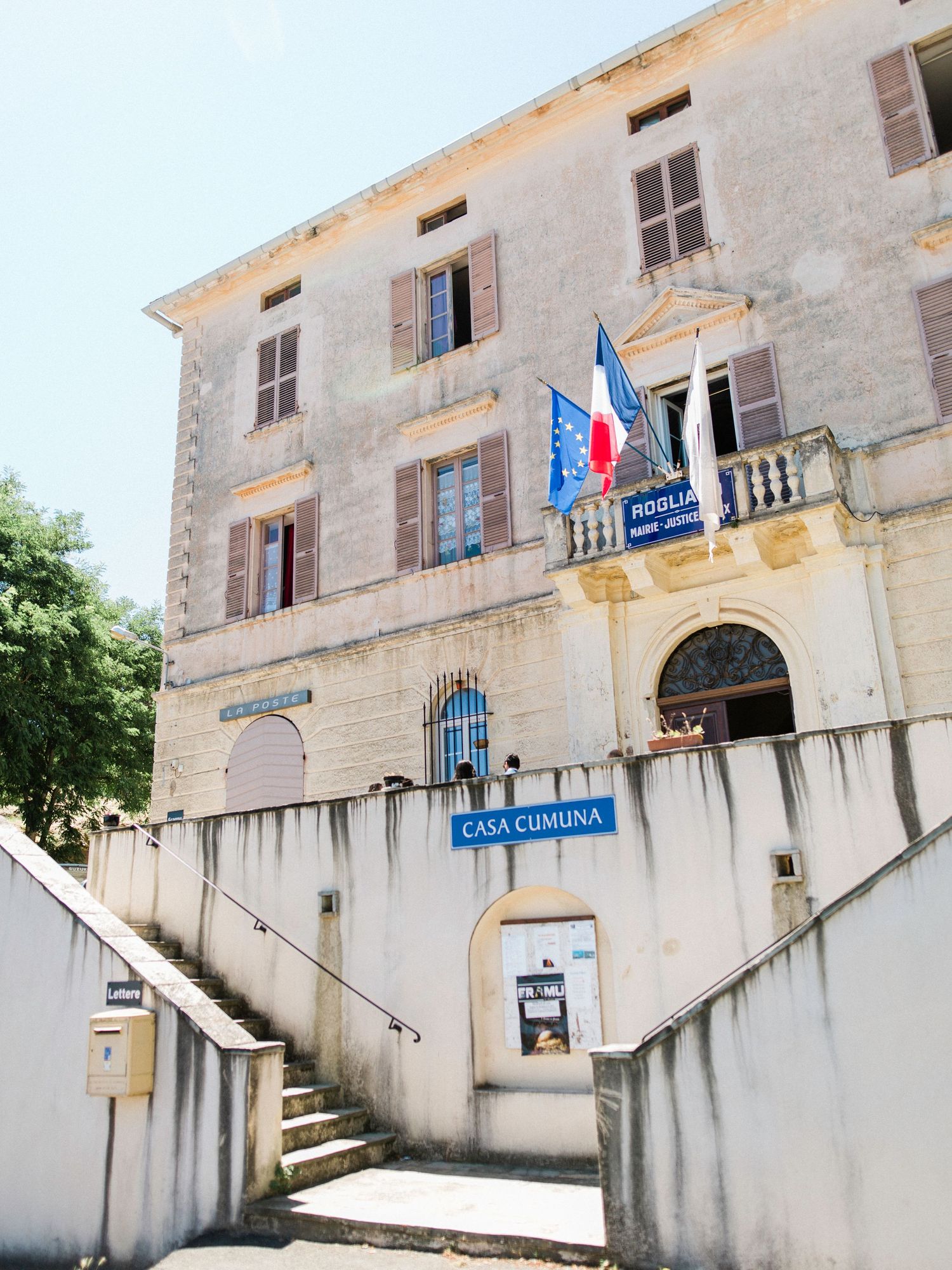 Historic Casa Cumuna building with French and EU flags, stone facade, balcony, and staircase entrance in Corsica, France.