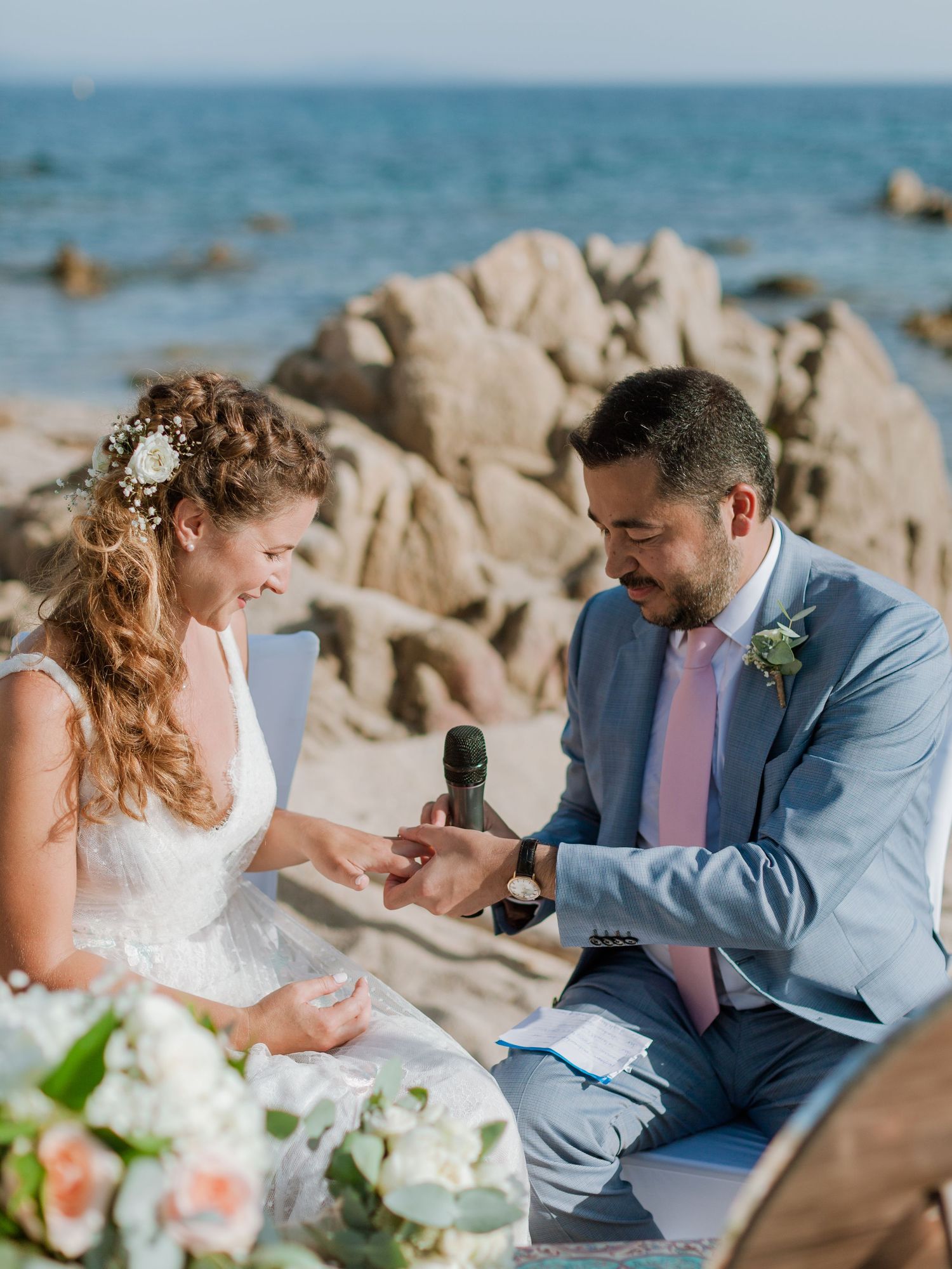 Le marié passe l'alliance au doigt de la mariée lors d'une cérémonie sur la plage, avec une côte rocheuse en arrière-plan.