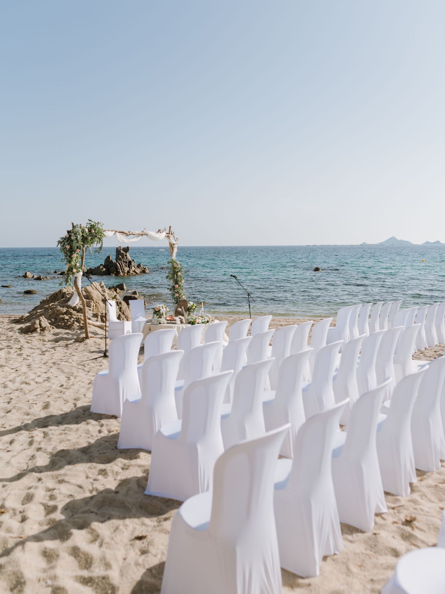 Installation pour une cérémonie de mariage sur la plage, avec des housses de chaise blanches disposées en rangées face à une arche fleurie