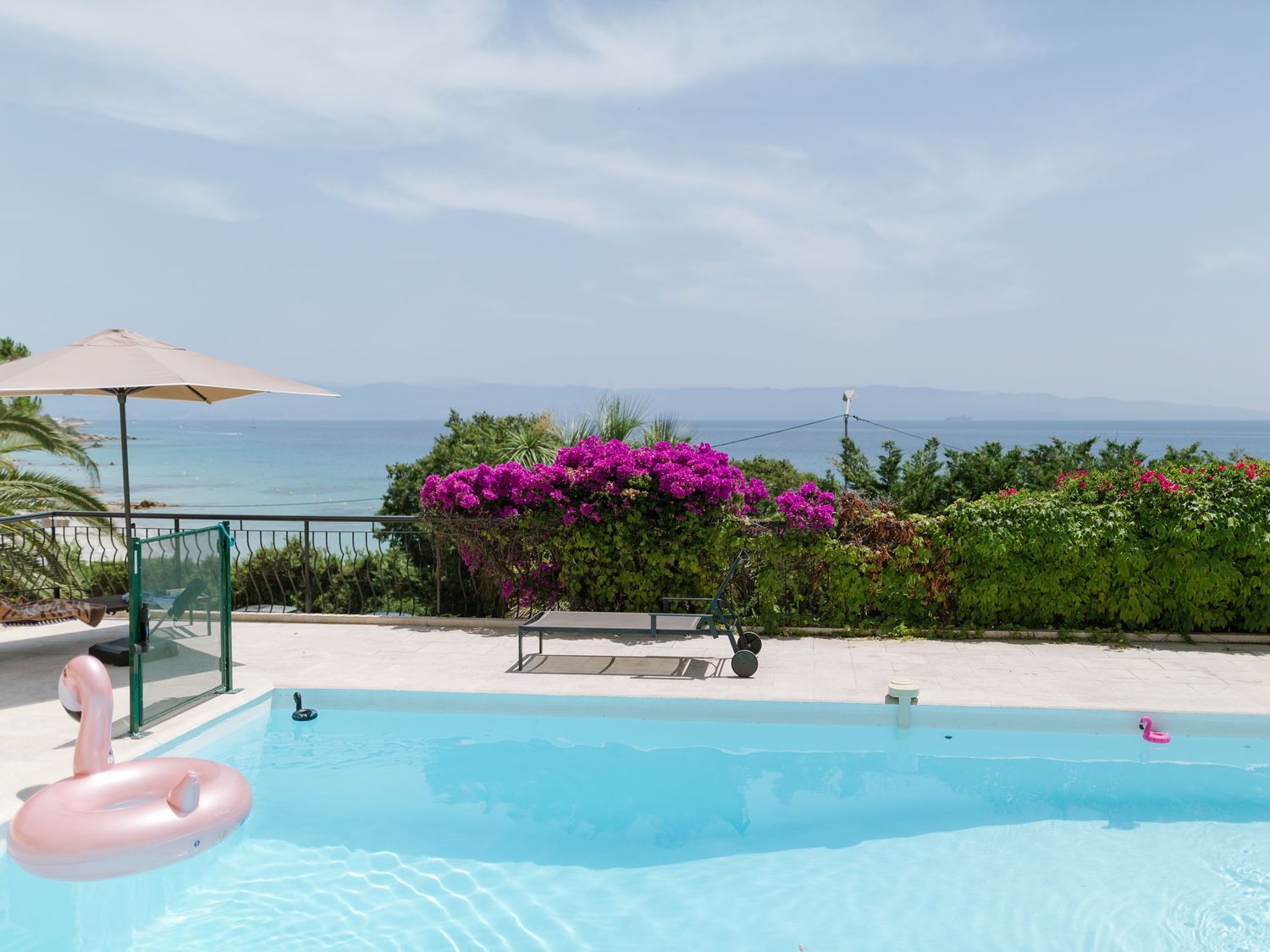 Piscine d'une villa de luxe avec une bouée flamant rose, des bougainvilliers et une vue imprenable sur la mer sous un ciel dégagé.