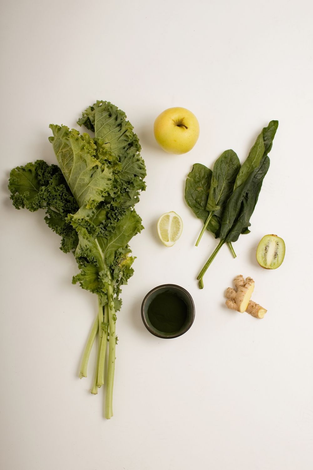 Vue à plat des ingrédients d'un smoothie vert, notamment du chou frisé, des épinards, de la pomme, du citron, du kiwi et du gingembre.
