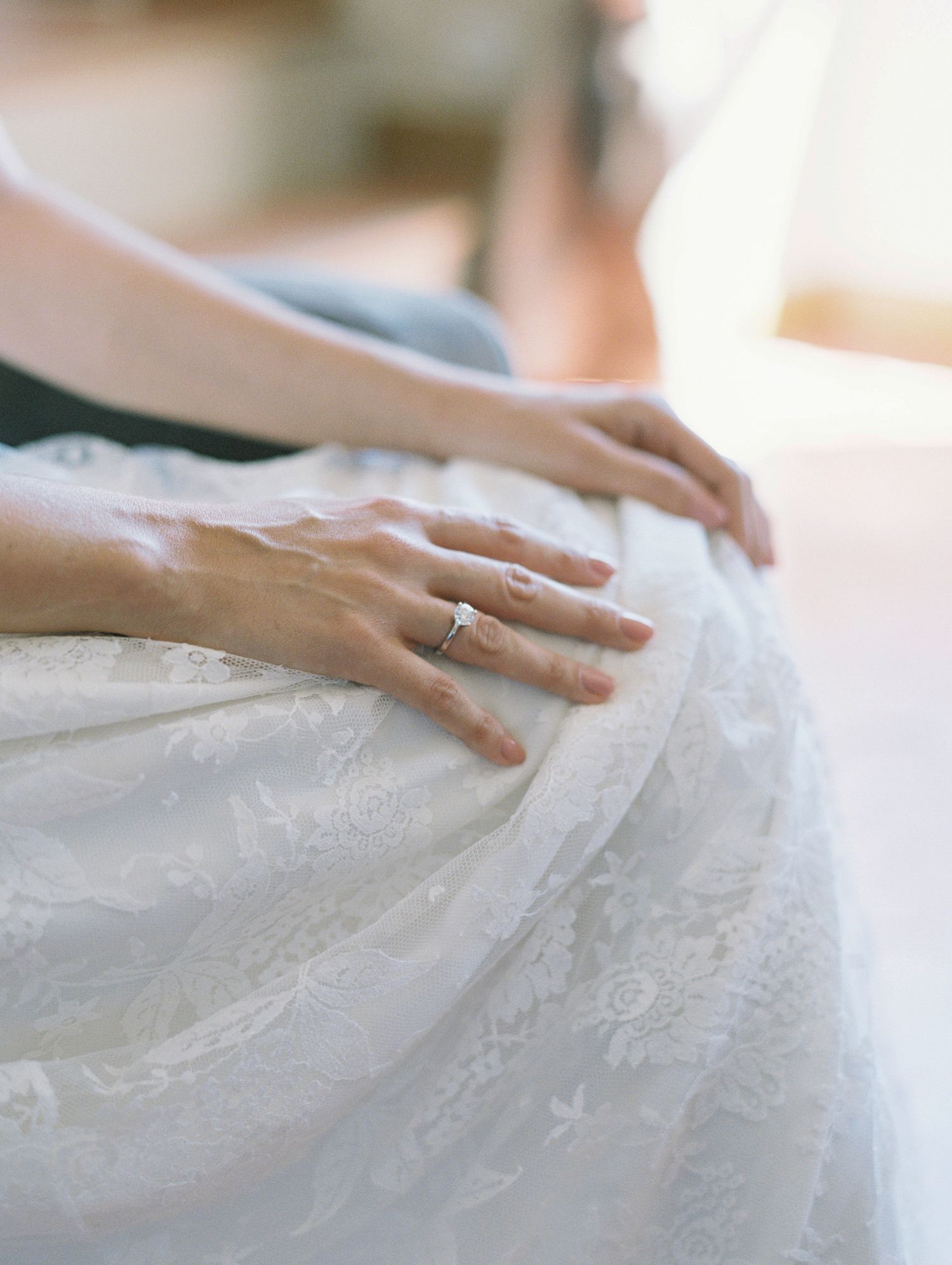 Close-up of a bride's hand with diamond engagement ring resting on white lace wedding dress fabric.
