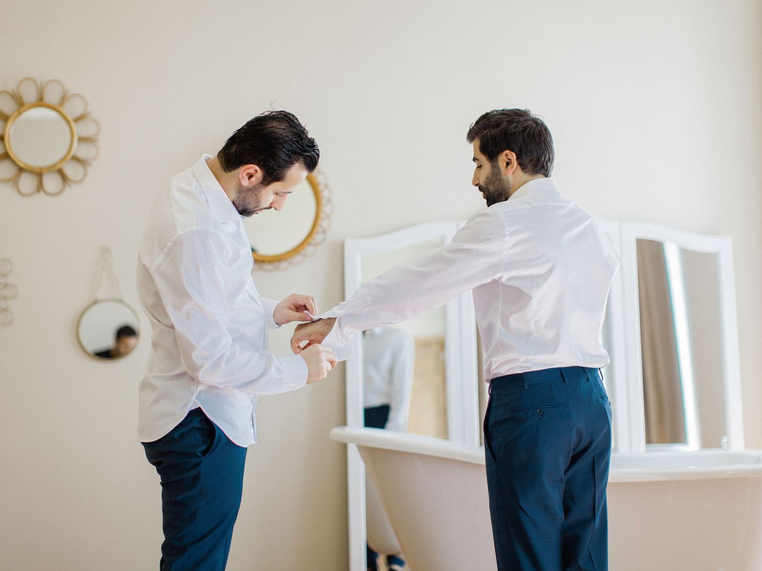 Two men in white shirts and navy trousers help each other with cufflinks before a formal event.