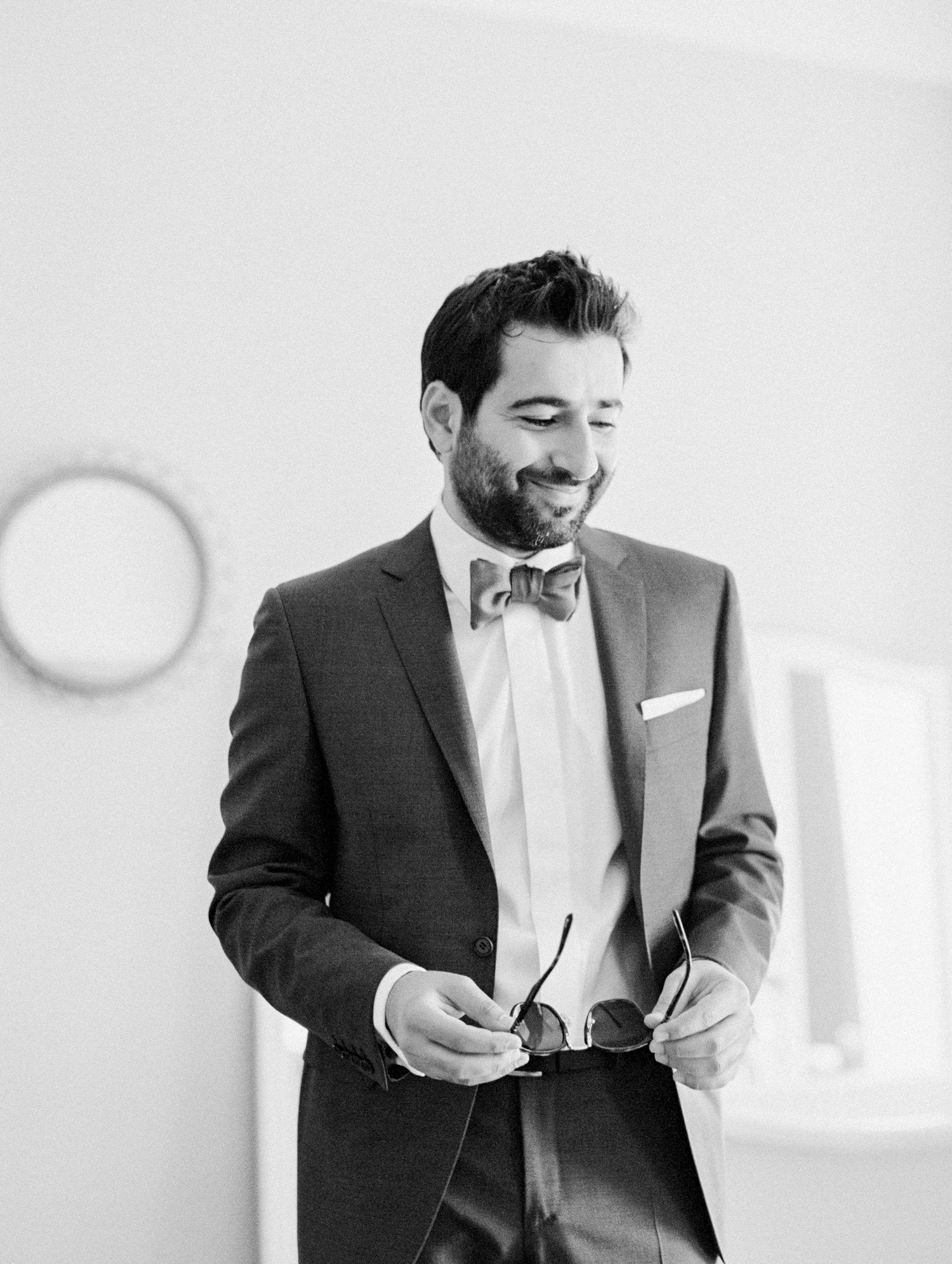 Smiling groom in a suit and bow tie holding sunglasses, black and white wedding portrait.