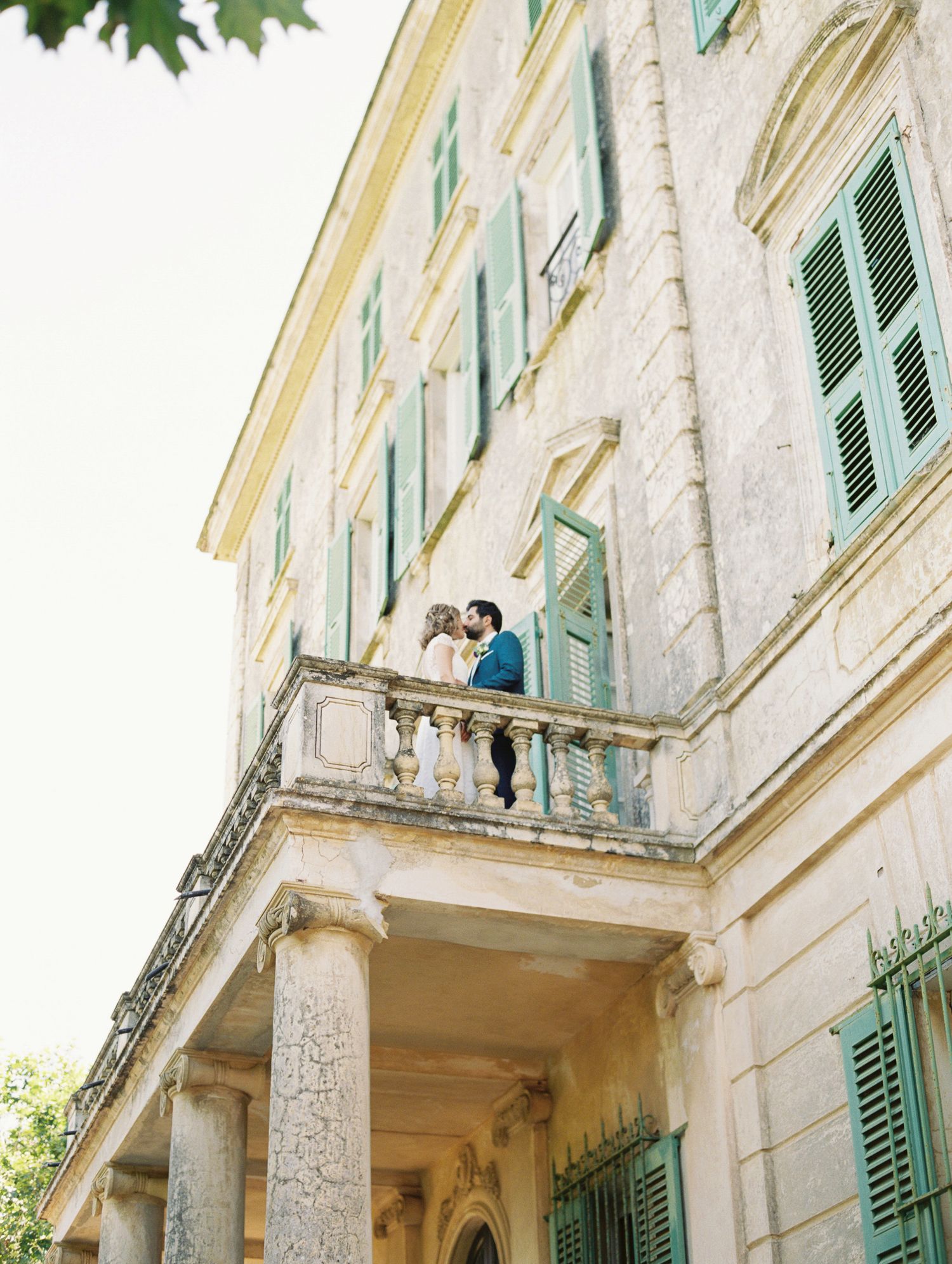 Couple sharing a romantic kiss on ornate balcony of elegant stone villa with green shutters.
