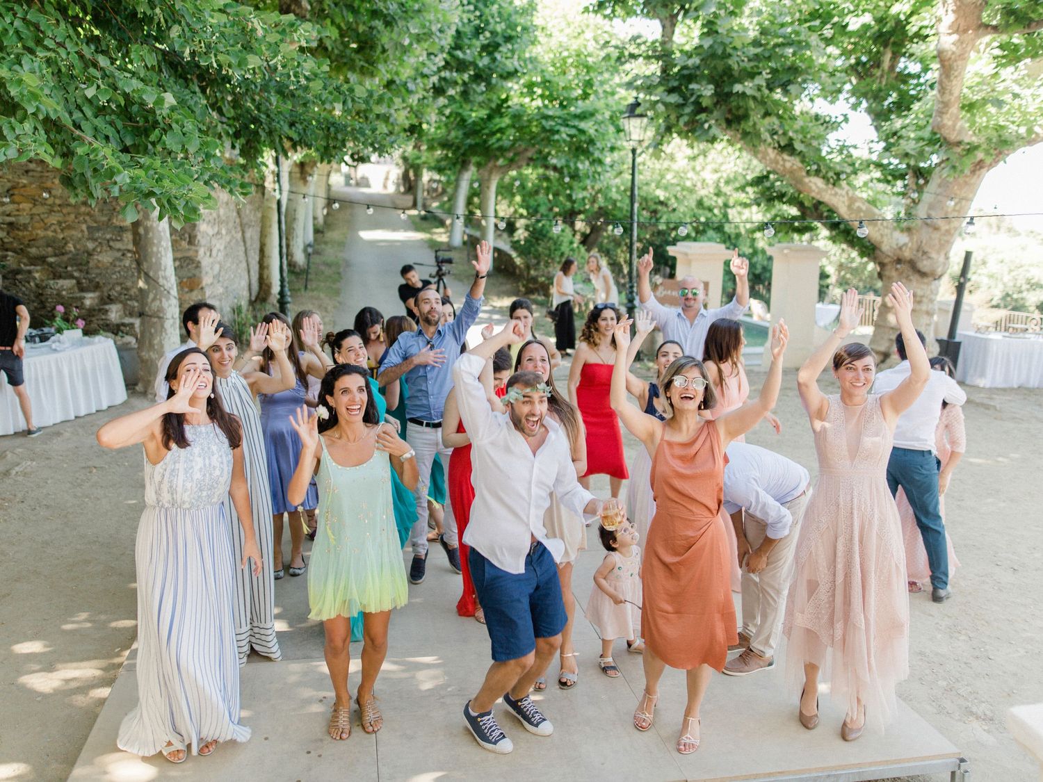Joyful wedding guests dancing and celebrating outdoors under green trees at a sunny summer wedding reception.
