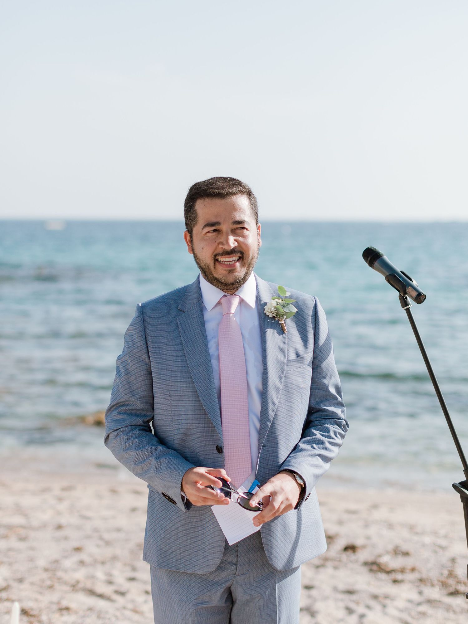 Marié souriant en costume gris, avec une cravate rose et une boutonnière, debout sur la plage lors d'une cérémonie de mariage en plein air.
