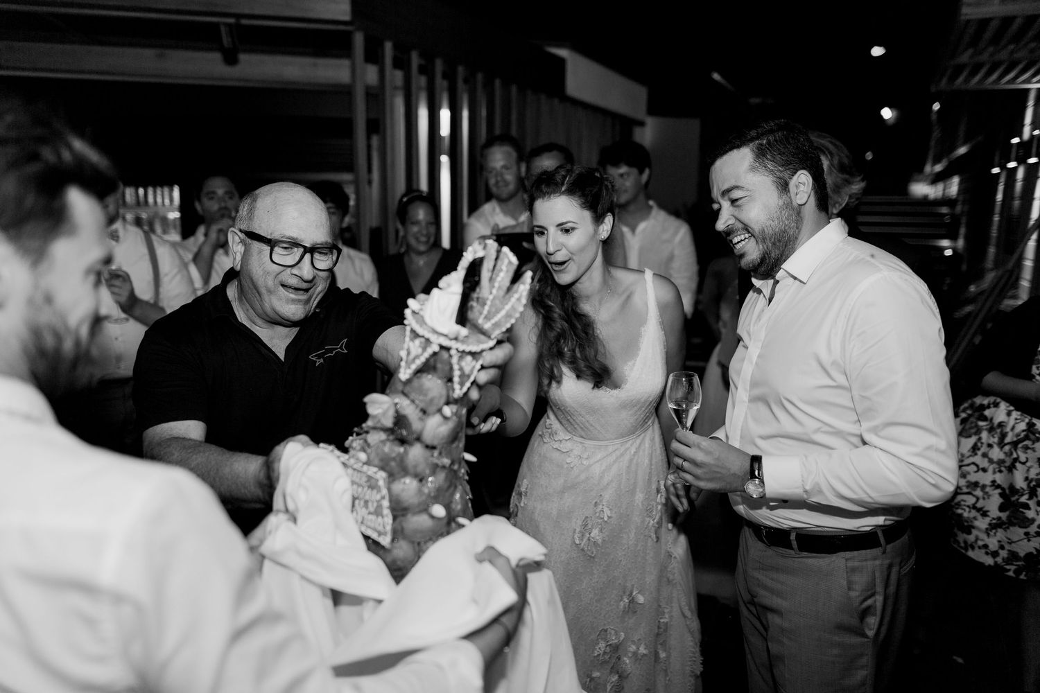 Photo en noir et blanc d'une mariée et d'un marié souriants à une table de réception de mariage, entourés d'invités.