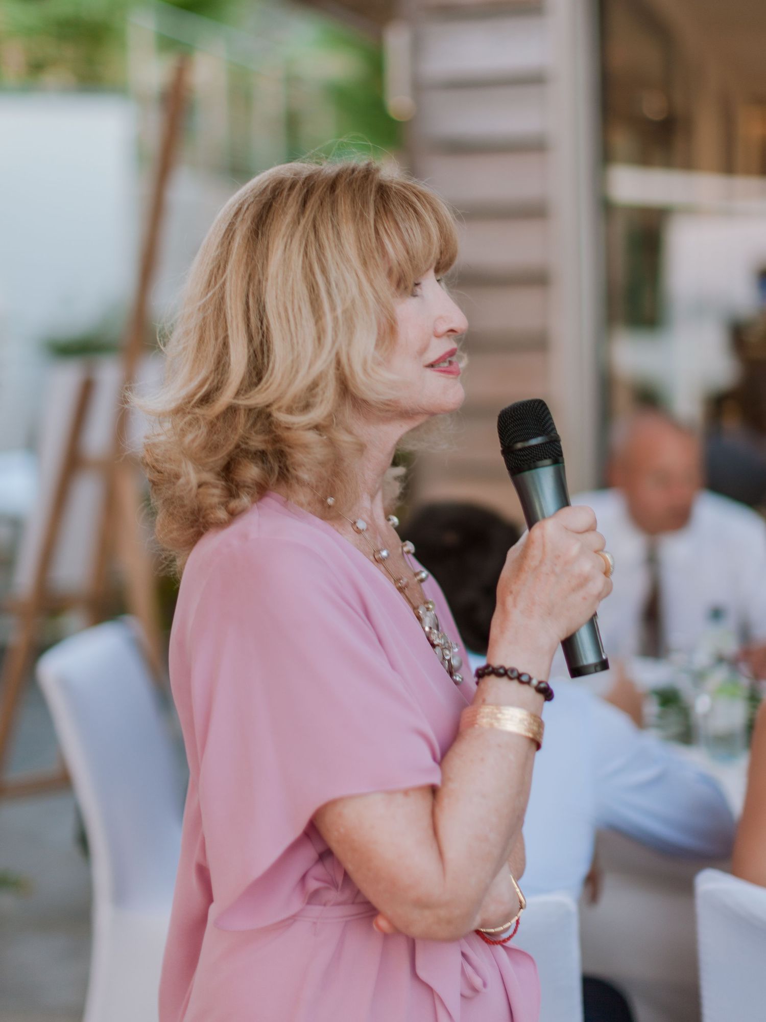 Une femme en robe rose parle dans un microphone lors d'un événement en plein air ; elle porte des bijoux et un bracelet.
