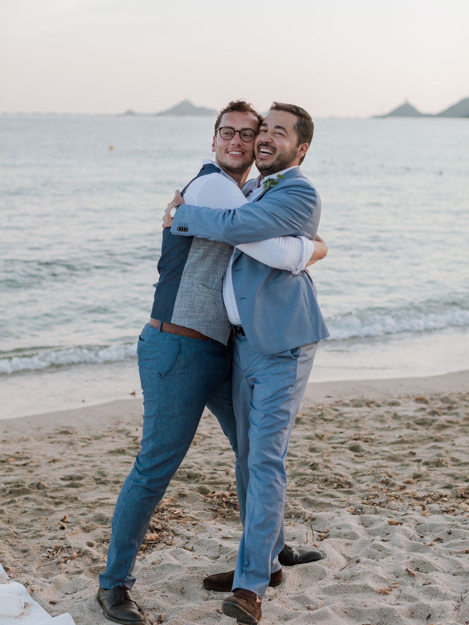 Deux hommes en costume bleu s'étreignent en souriant sur une plage de sable au coucher du soleil, avec les vagues de l'océan en arrière-plan