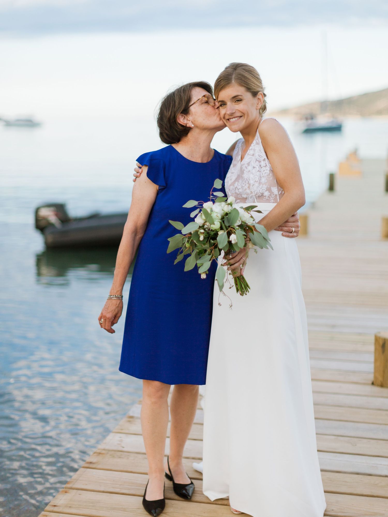Une mariée en robe blanche tient son bouquet tandis qu'une femme plus âgée en robe bleue l'embrasse sur la joue