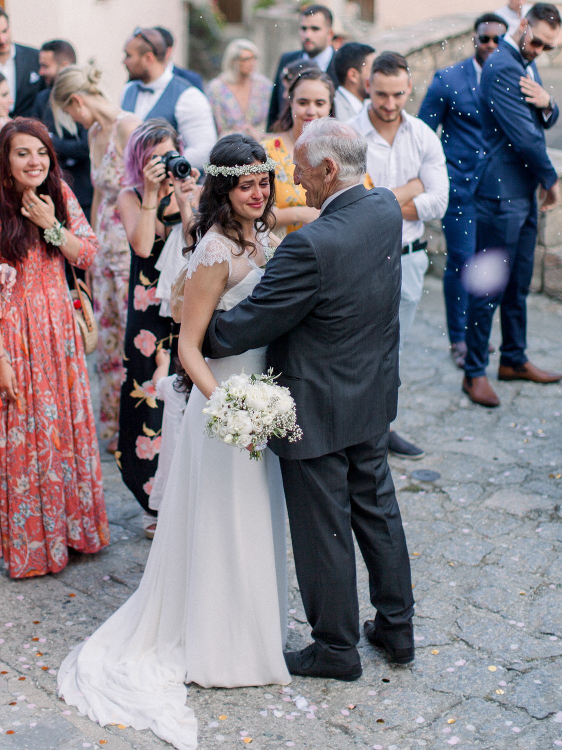 Bride in white gown with floral crown embraces elderly man at outdoor wedding celebration with guests.