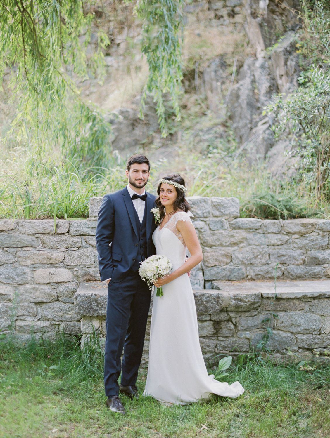 Bride and groom posing together outdoors near a stone wall, she holds a white bouquet, he wears a navy suit with bow tie.