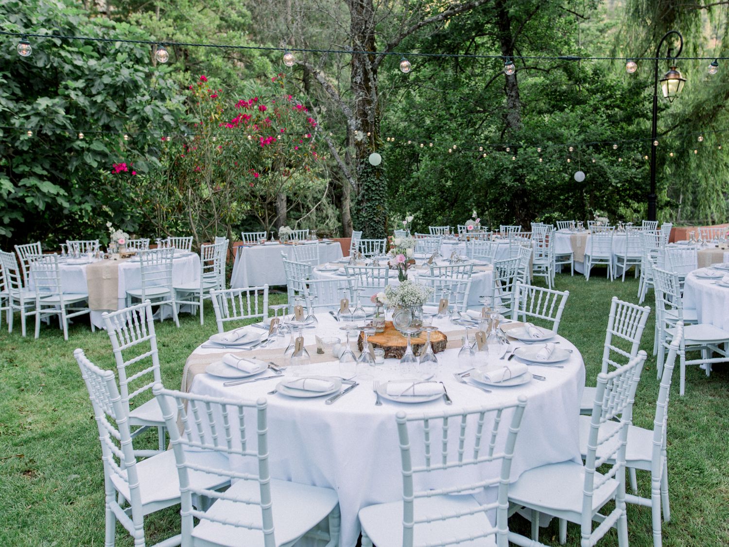 Elegant outdoor wedding reception with white Chiavari chairs and round tables set on a lush green lawn surrounded by trees.